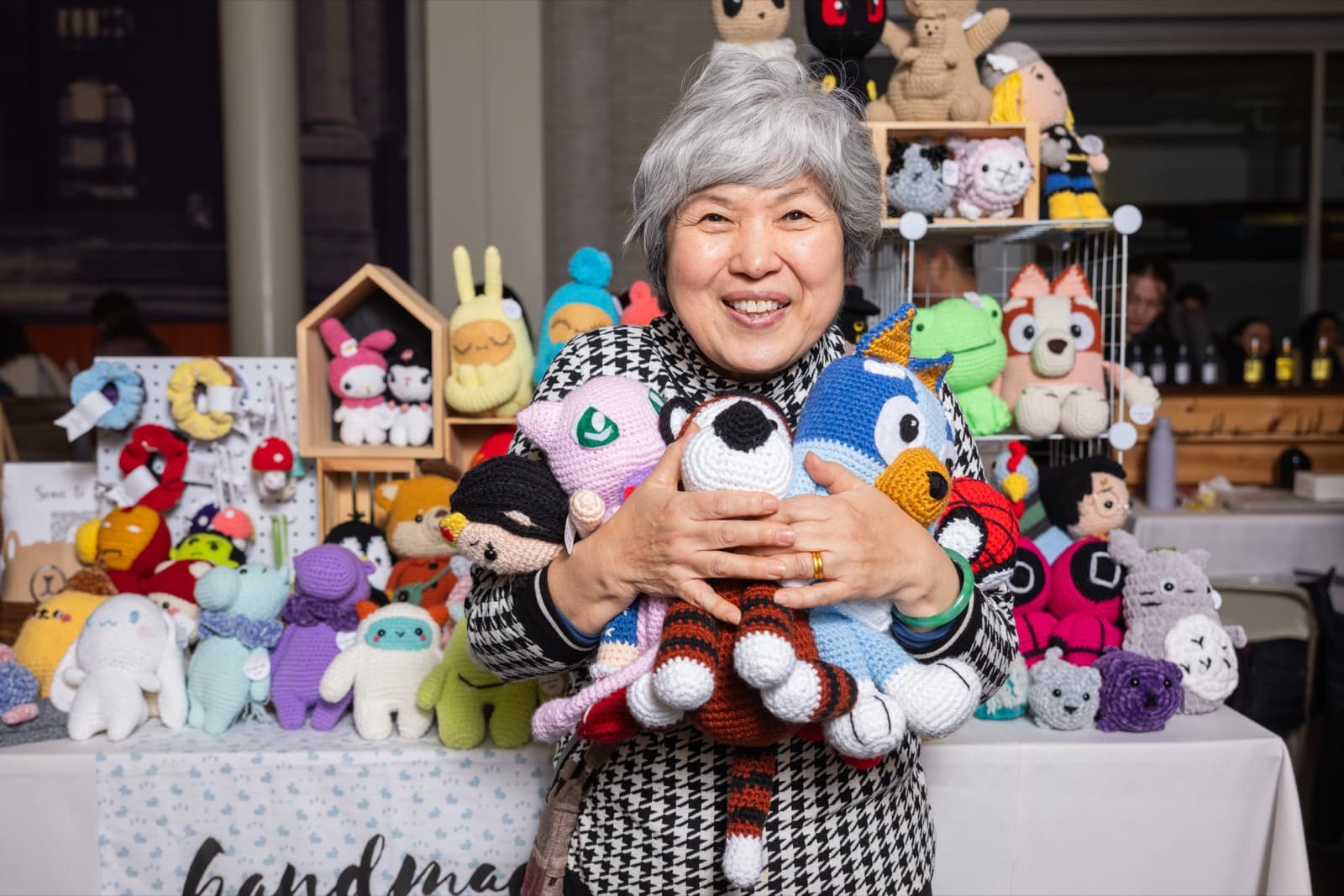 Woman smiling while holding an armful of colorful handmade crochet toys at a craft fair table.