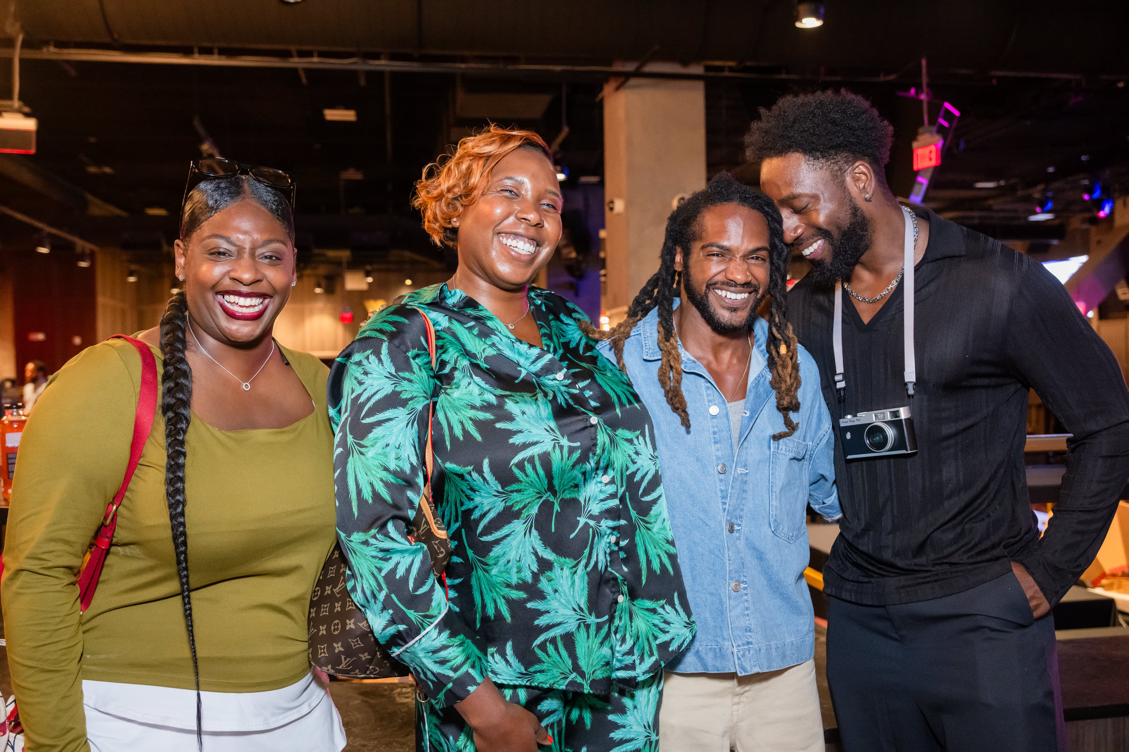 Four friends laughing together at an indoor venue, one holding a camera around his neck.