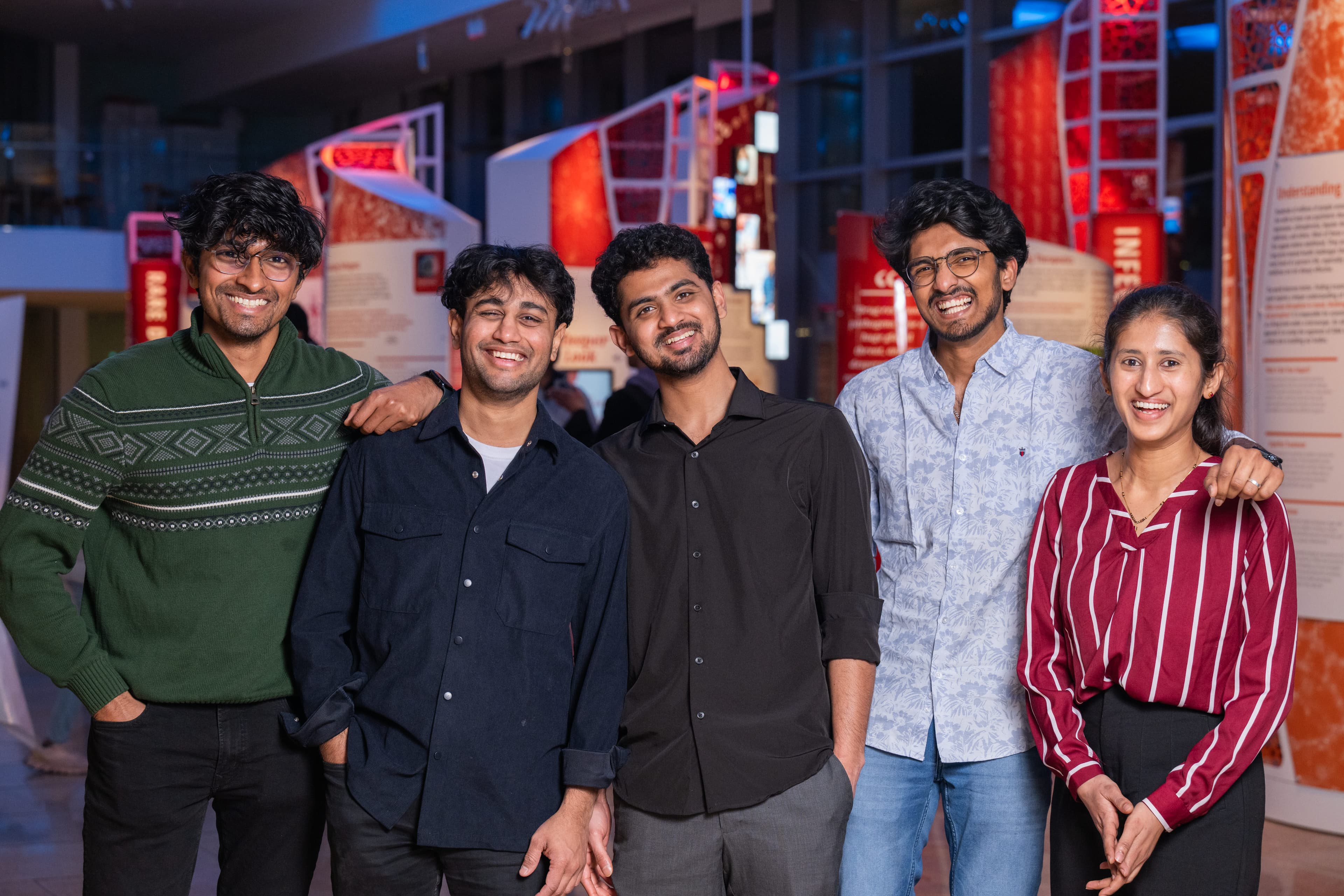 Five friends posing together arm-in-arm at an exhibit hall, laughing, illuminated red panels behind them.