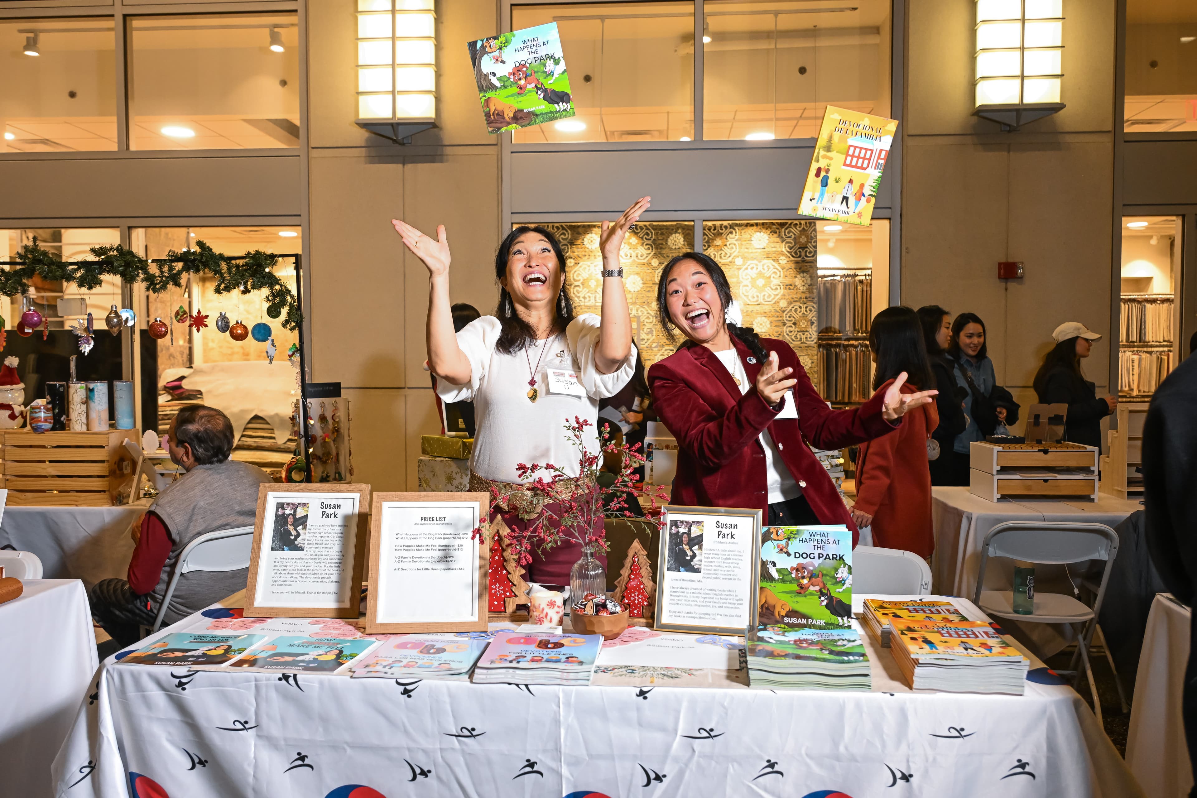 Two women throwing children's books into the air, laughing, at a holiday vendor table.
