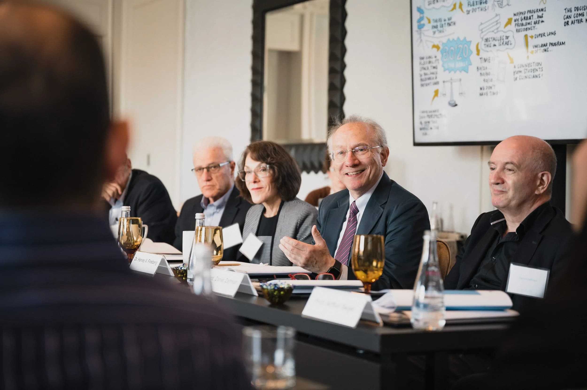 Group of professionals seated around a conference table, one man gesturing while speaking, a whiteboard with notes behind them.
