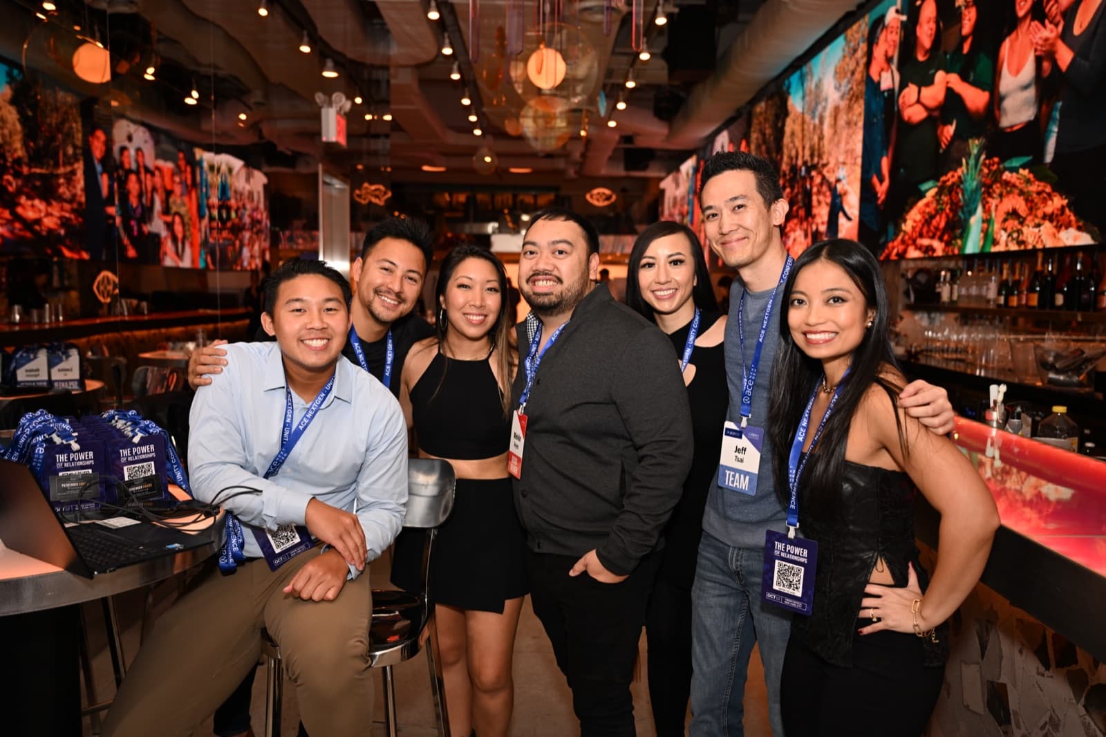 Seven people posed together smiling at a lounge venue, string lights and colorful wall art behind them.