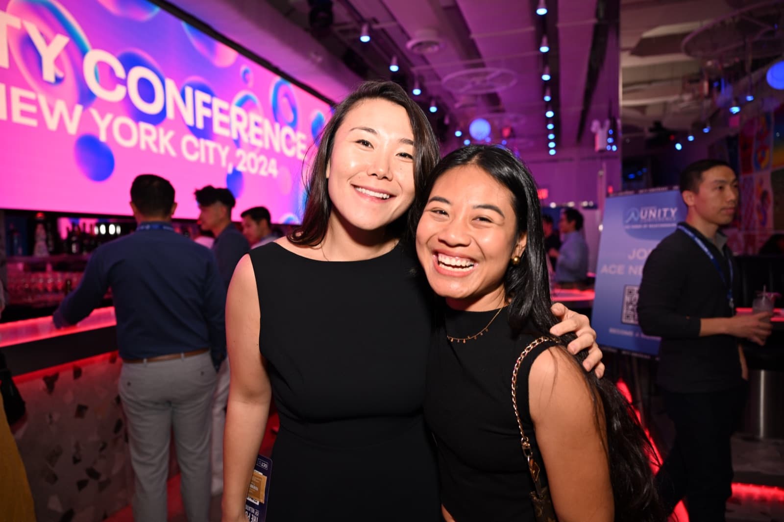 Two women embracing and smiling, a large screen with event branding behind them, a bar visible nearby.
