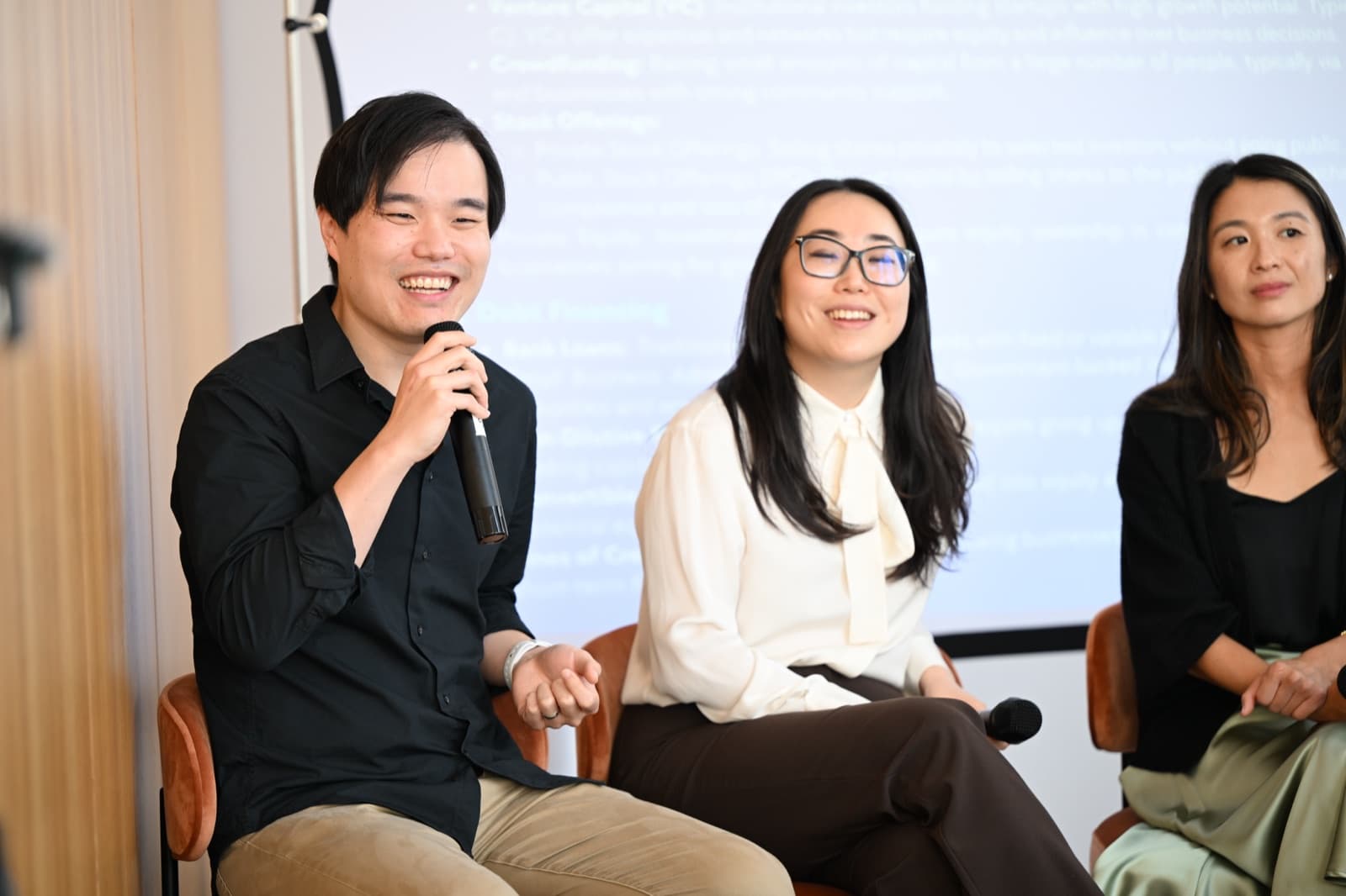 Man speaking into a microphone, grinning broadly, two women seated next to him, a presentation screen behind them.