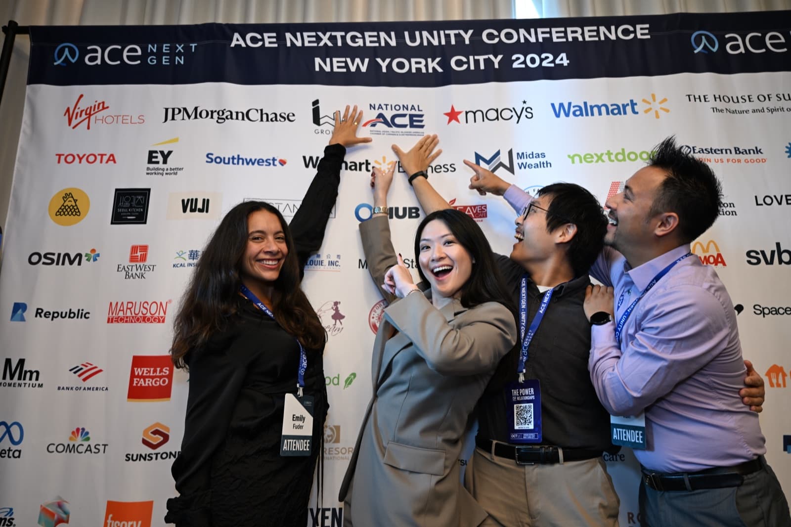 Four people cheering and pointing at a sponsor-logo backdrop, laughing together.