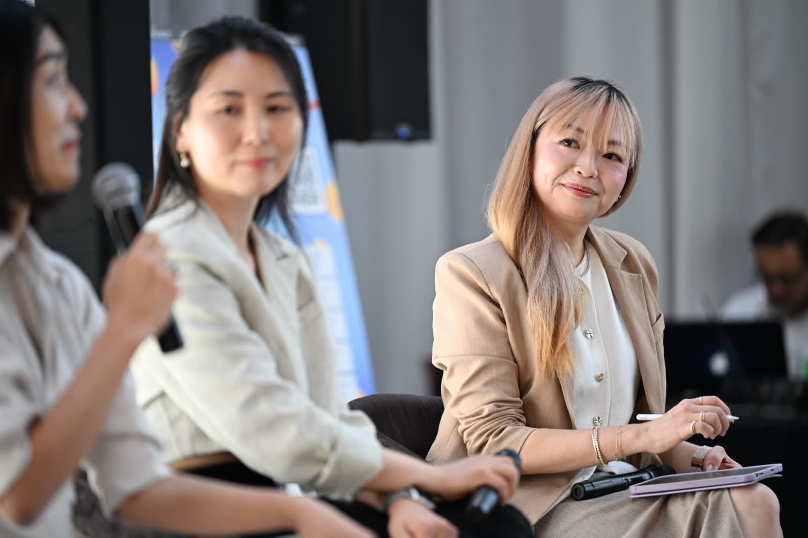 Woman in a tan blazer smiling and listening, two other people partially visible beside her.