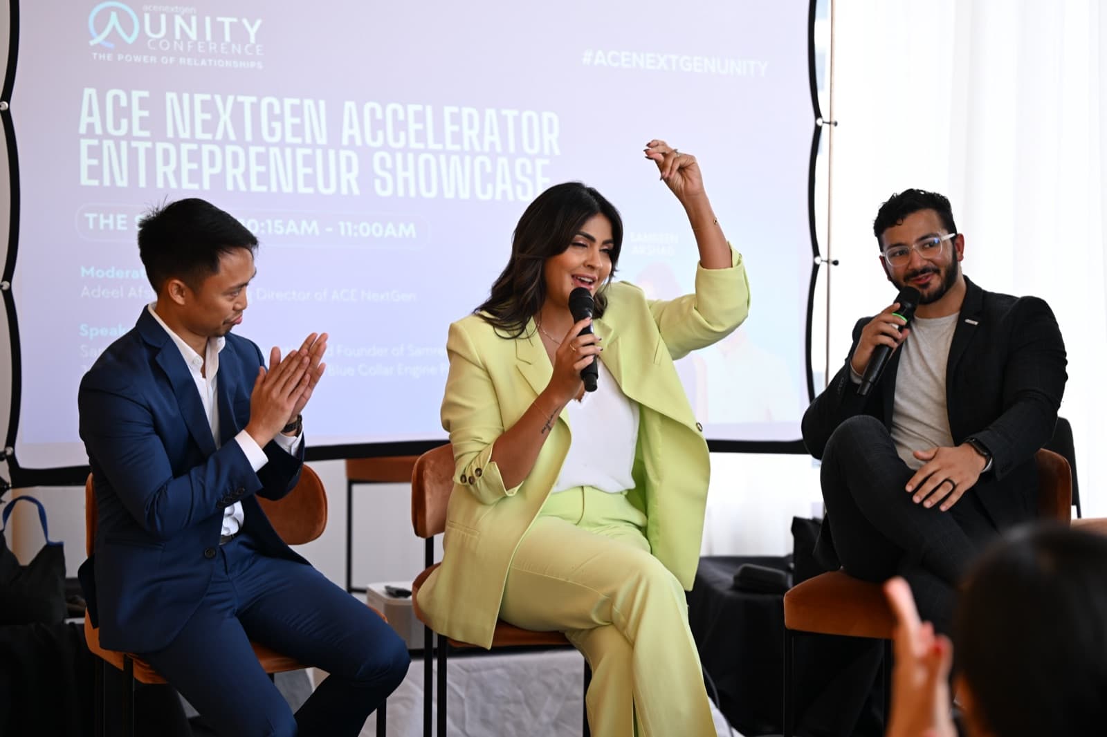 Woman gesturing while speaking into a microphone, two men seated beside her also holding microphones.
