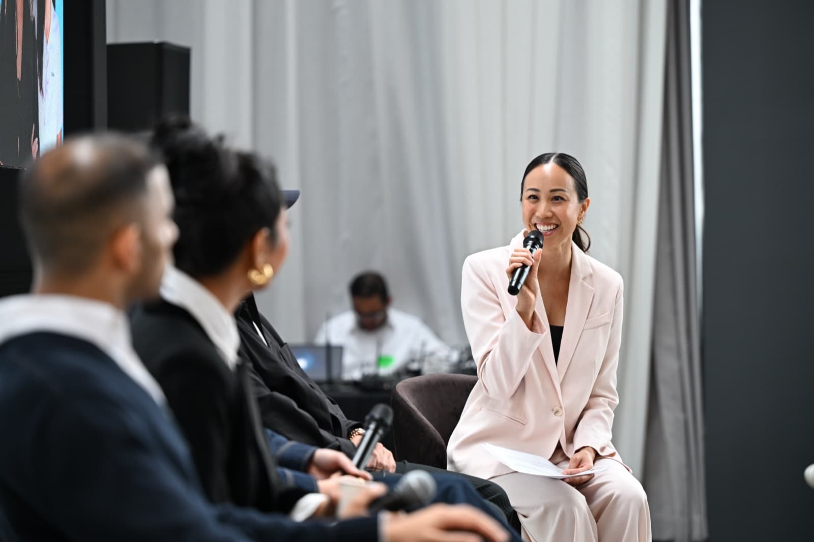 Woman speaking into a microphone, three other panelists seated nearby, a string curtain backdrop.