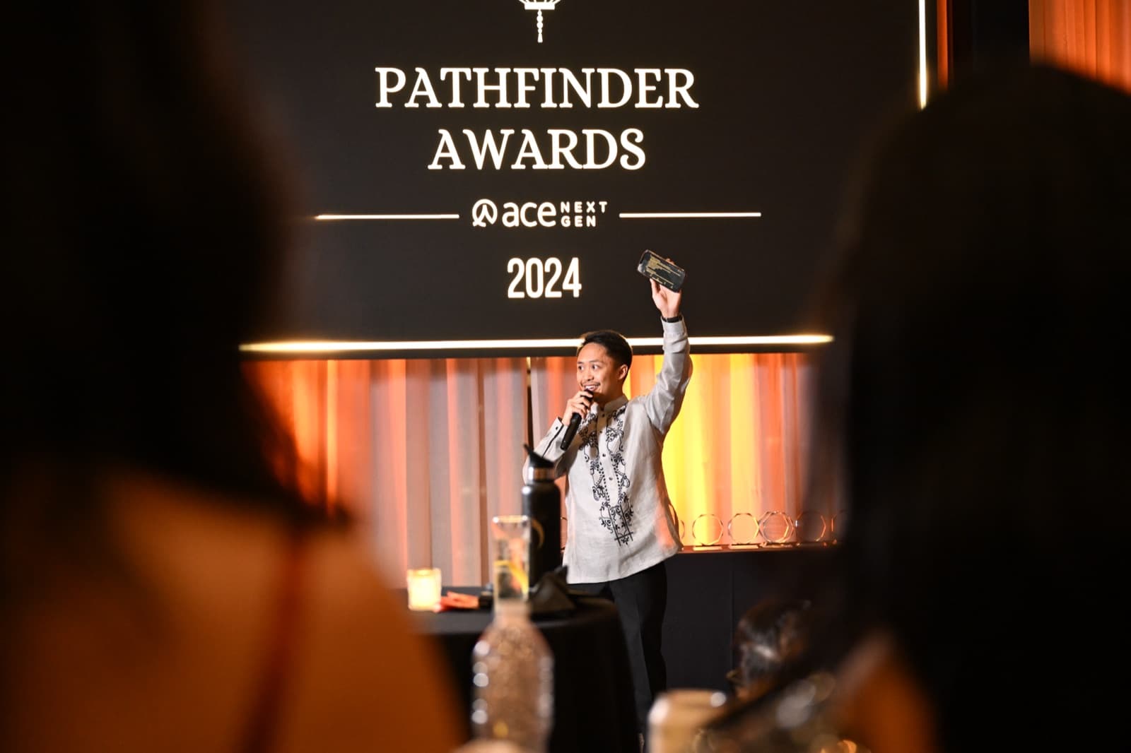 Man on stage holding an award plaque up triumphantly, an awards-ceremony backdrop and trophies behind him.