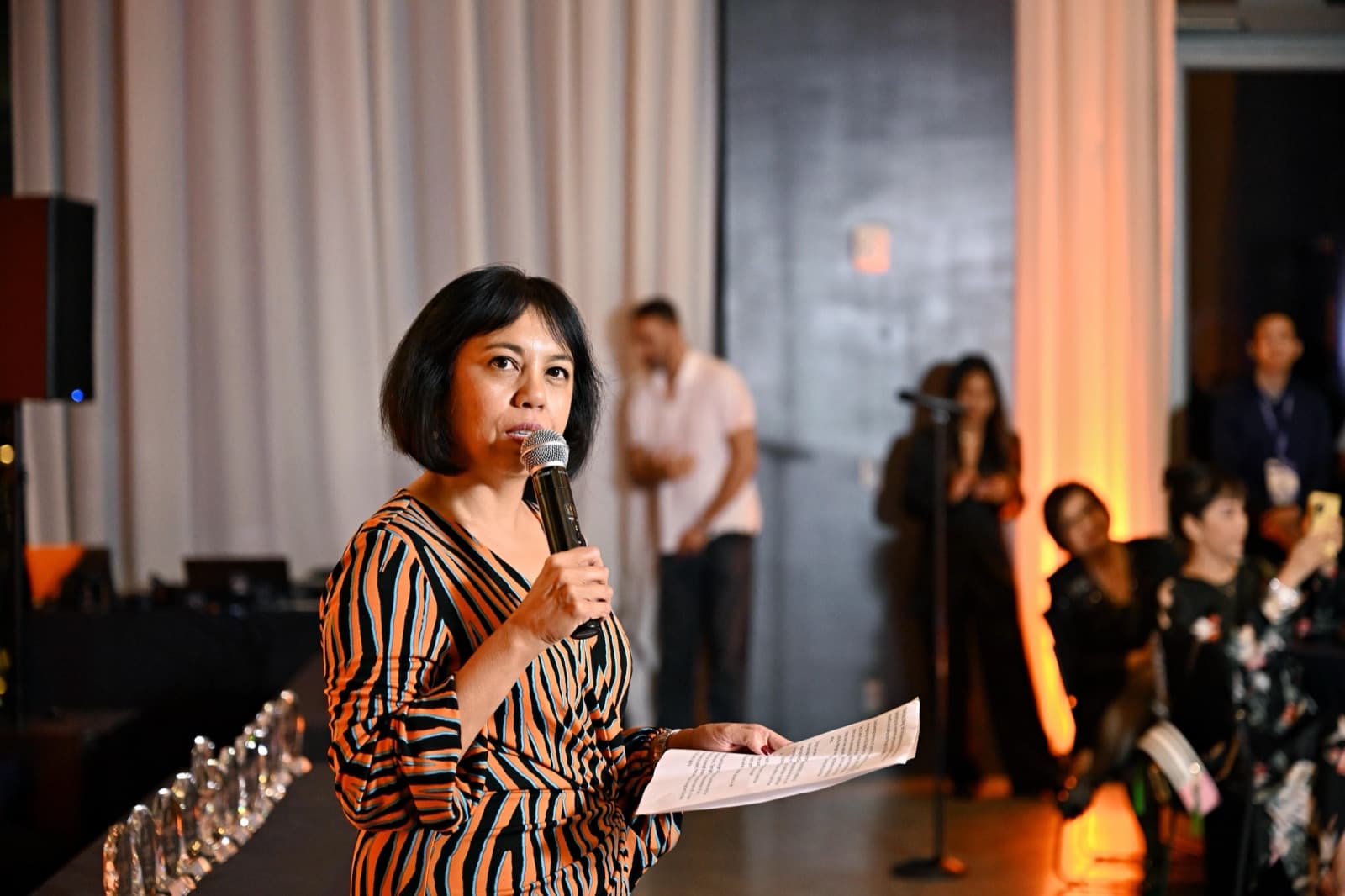 Woman speaking into a microphone, holding a paper, trophies lined up on a table beside her.