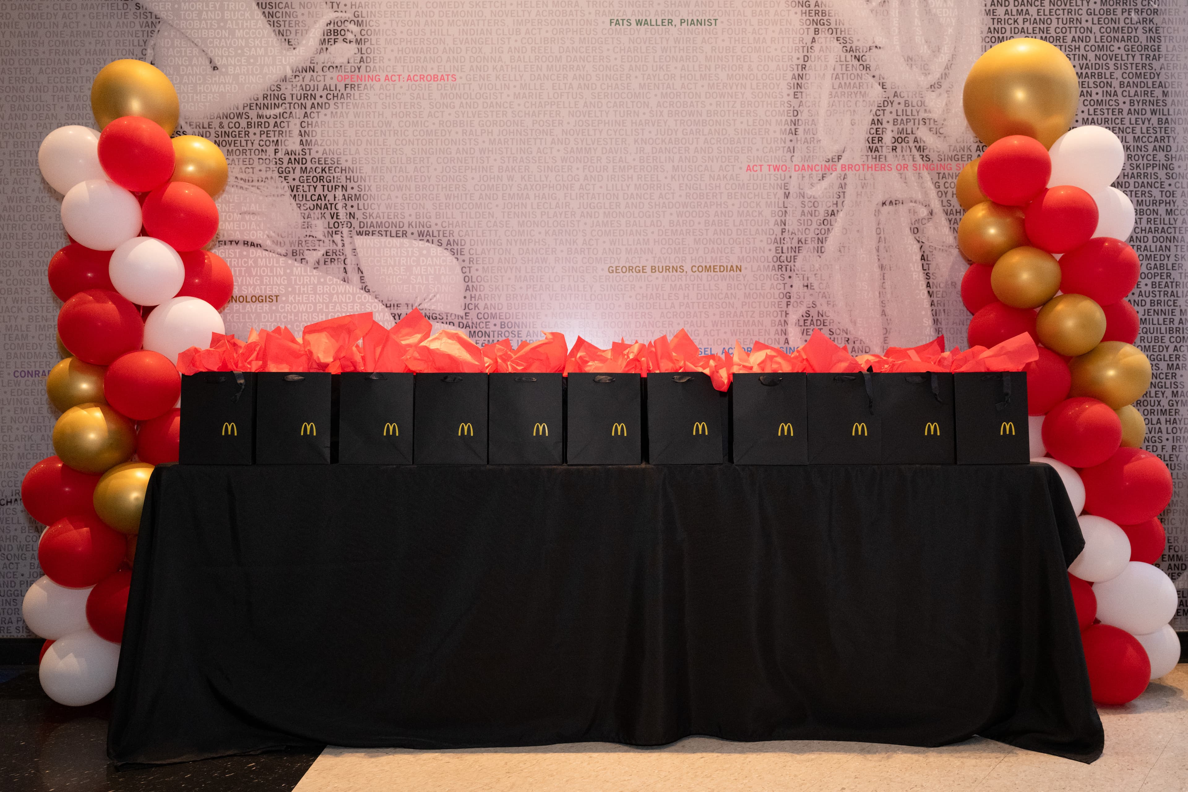 A row of black McDonald's gift bags on a skirted table, flanked by red, white, and gold balloon garlands.