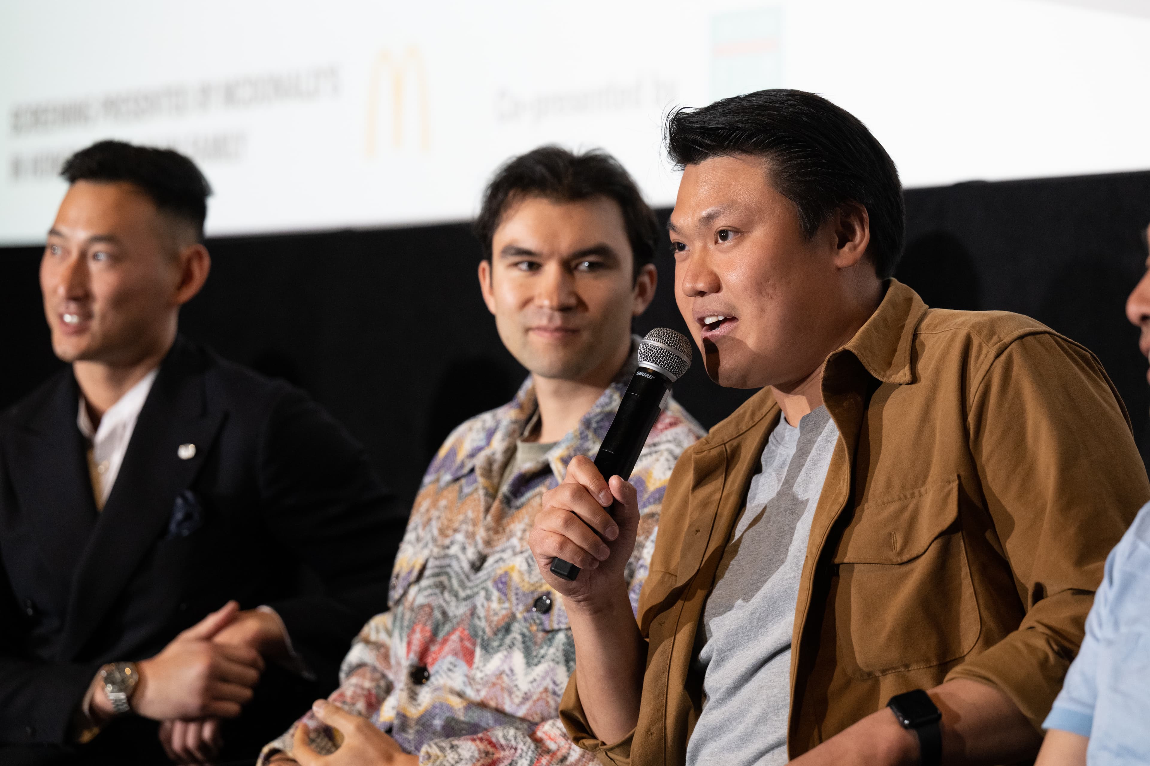 Three men seated on stage for a panel discussion, one speaking into a handheld microphone mid-sentence.