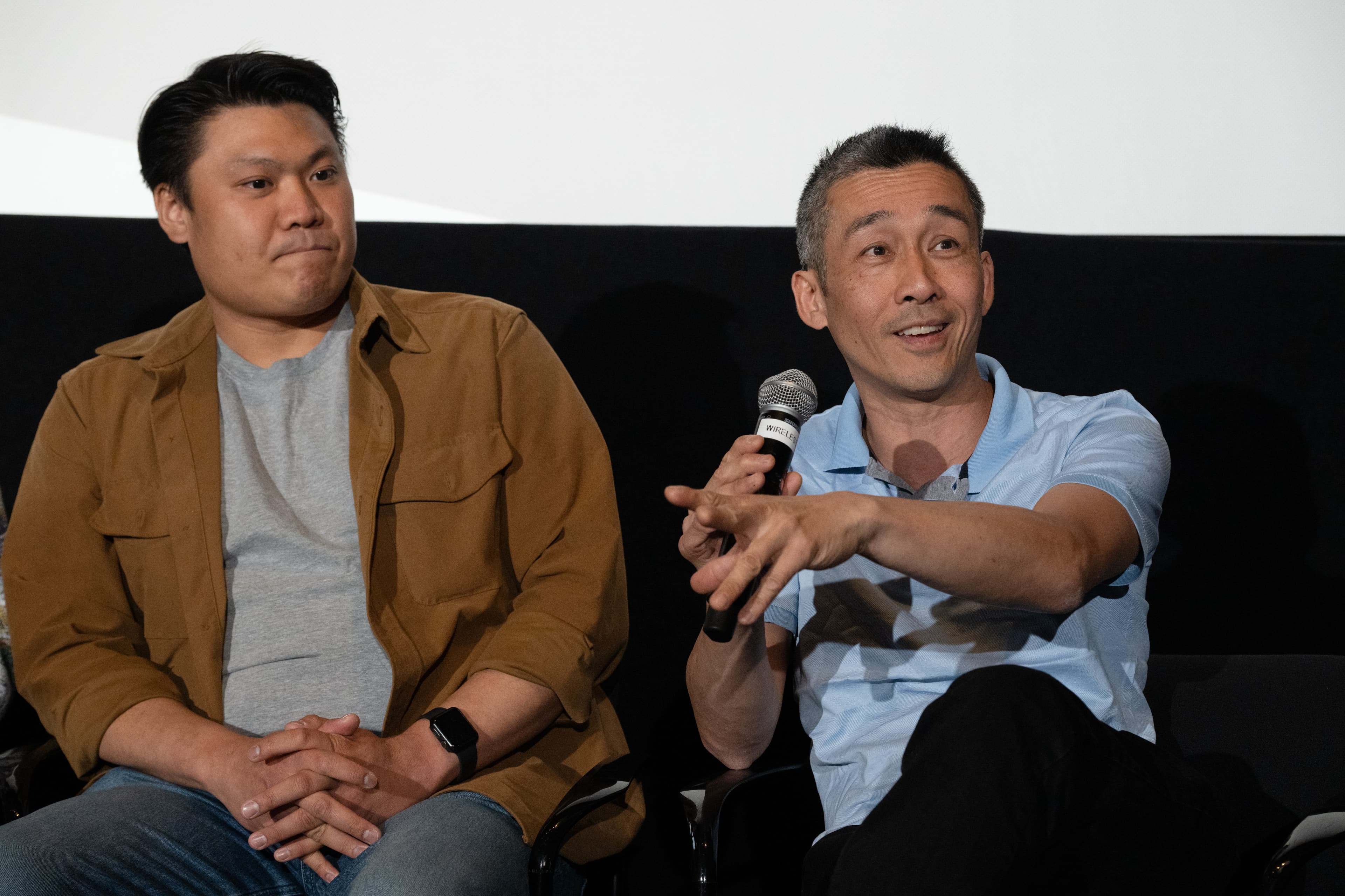 Two men seated on stage for a panel discussion, one gesturing while speaking into a handheld microphone.