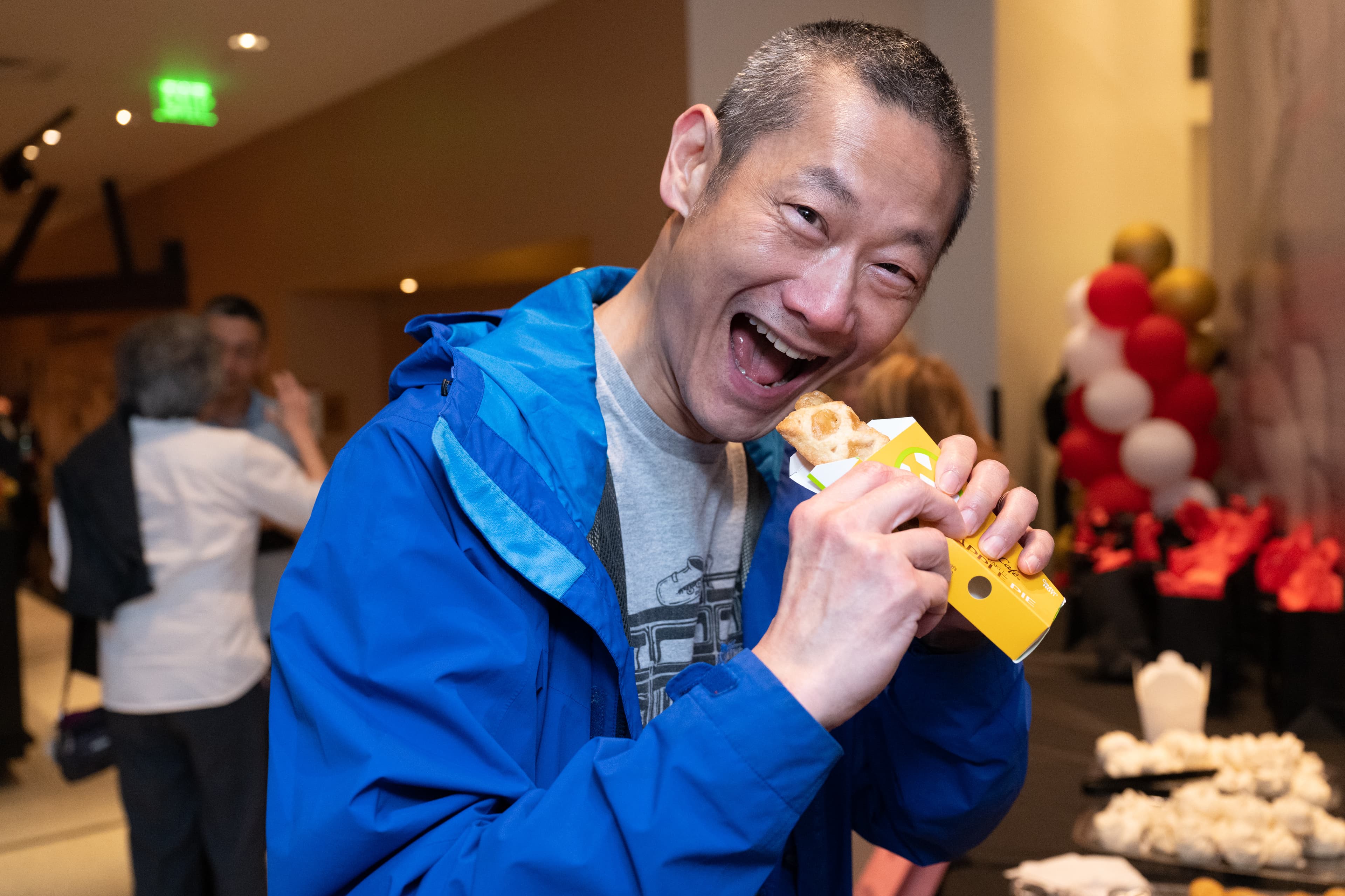 Man in a blue jacket laughing while holding a small pastry box, red and gold balloons visible behind him.