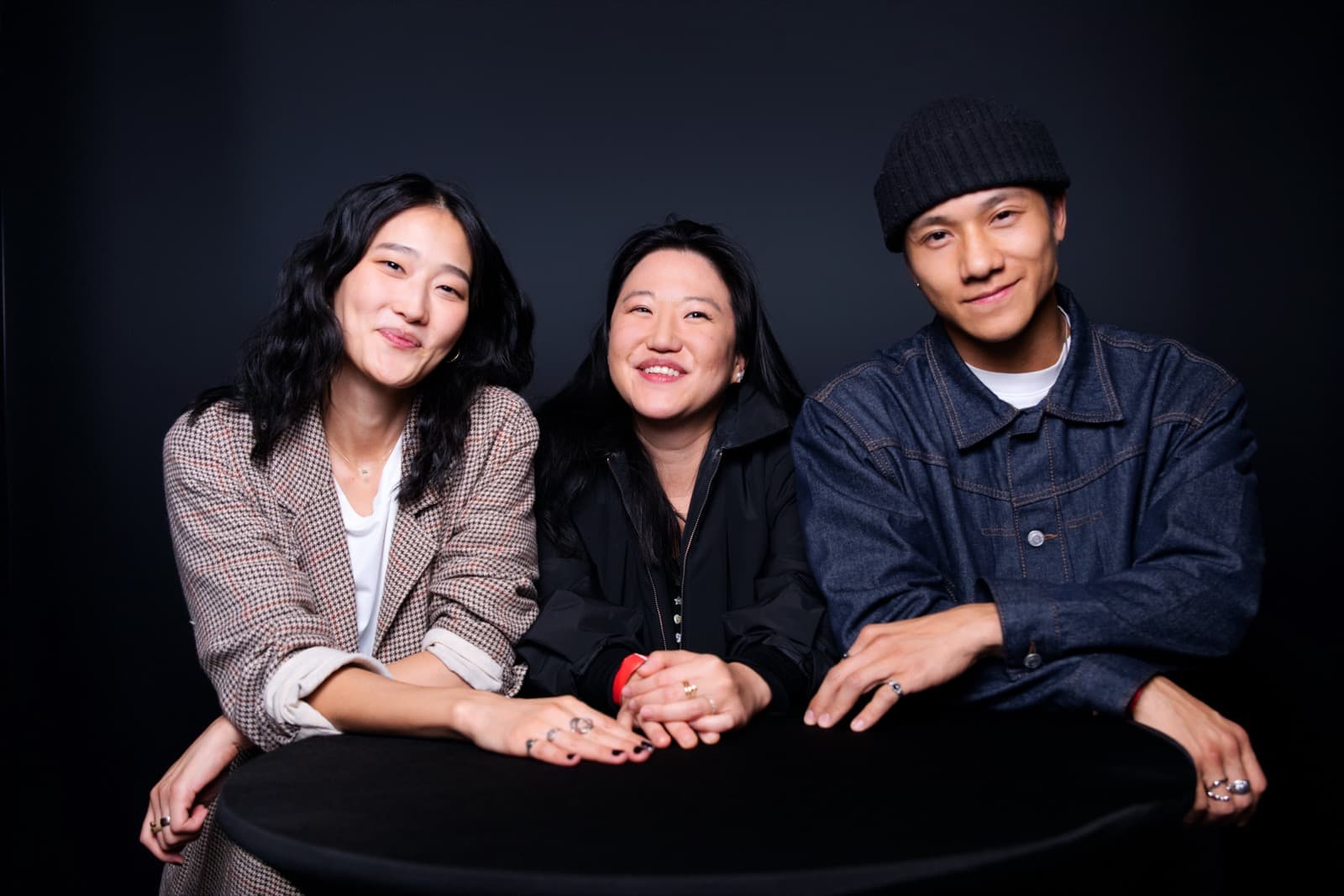 Three people leaning on a round table in a studio portrait, one in a houndstooth blazer, smiling at the camera.