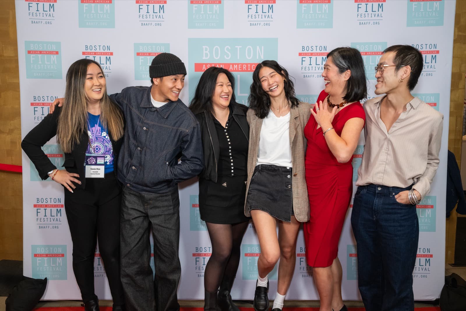 Six people laughing together at a step-and-repeat photo wall.