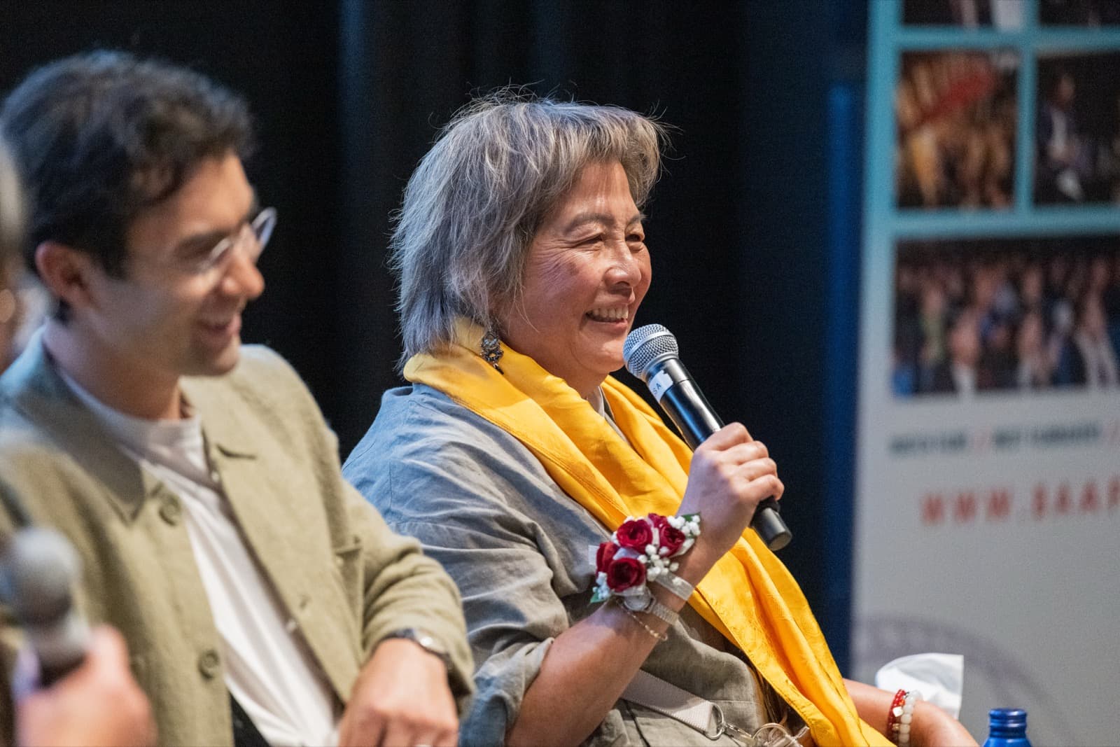 Woman with a yellow scarf and a red rose corsage grinning into a microphone on stage, a man smiling beside her.