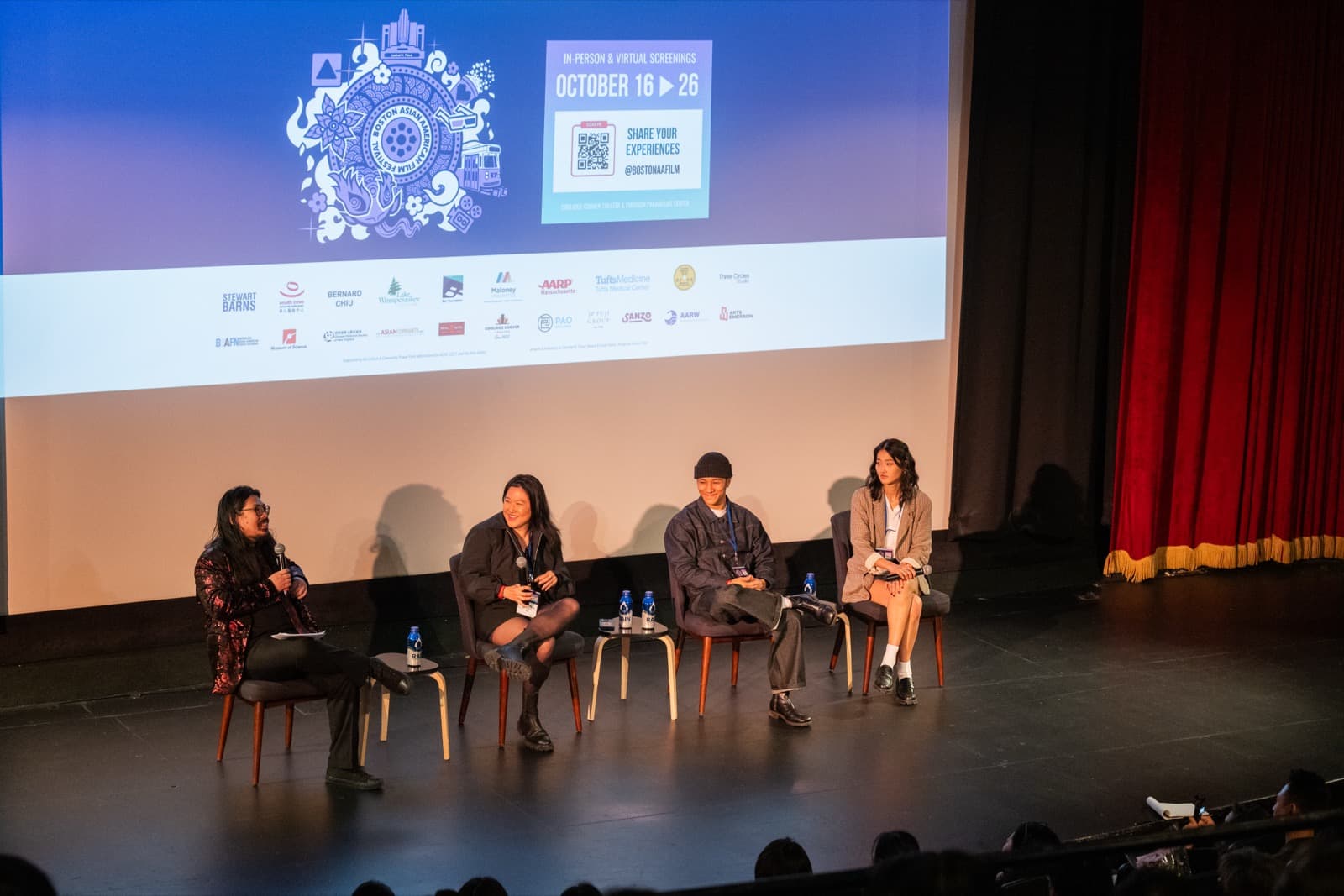 Four panelists seated on stage below a screen reading “In-Person & Virtual Screenings, October 16–26,” a red curtain behind them.