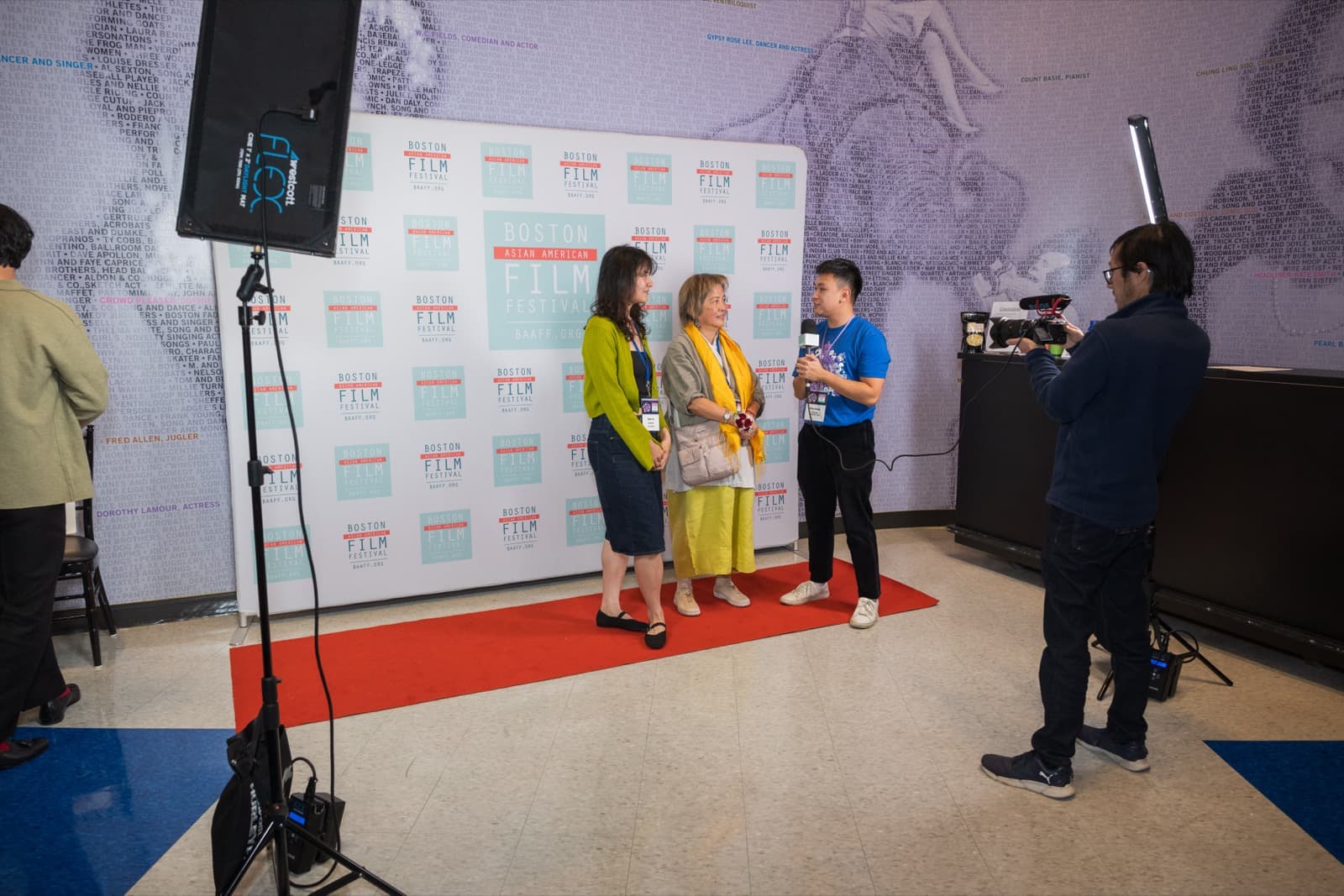 Two women being interviewed on a red carpet in front of a step-and-repeat, a videographer filming from the side.