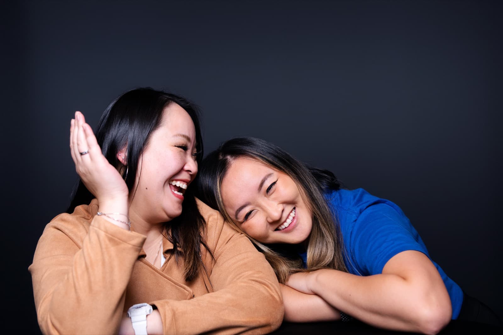 Two women laughing cheek to cheek, one in a tan jacket, arms crossed in a studio portrait.