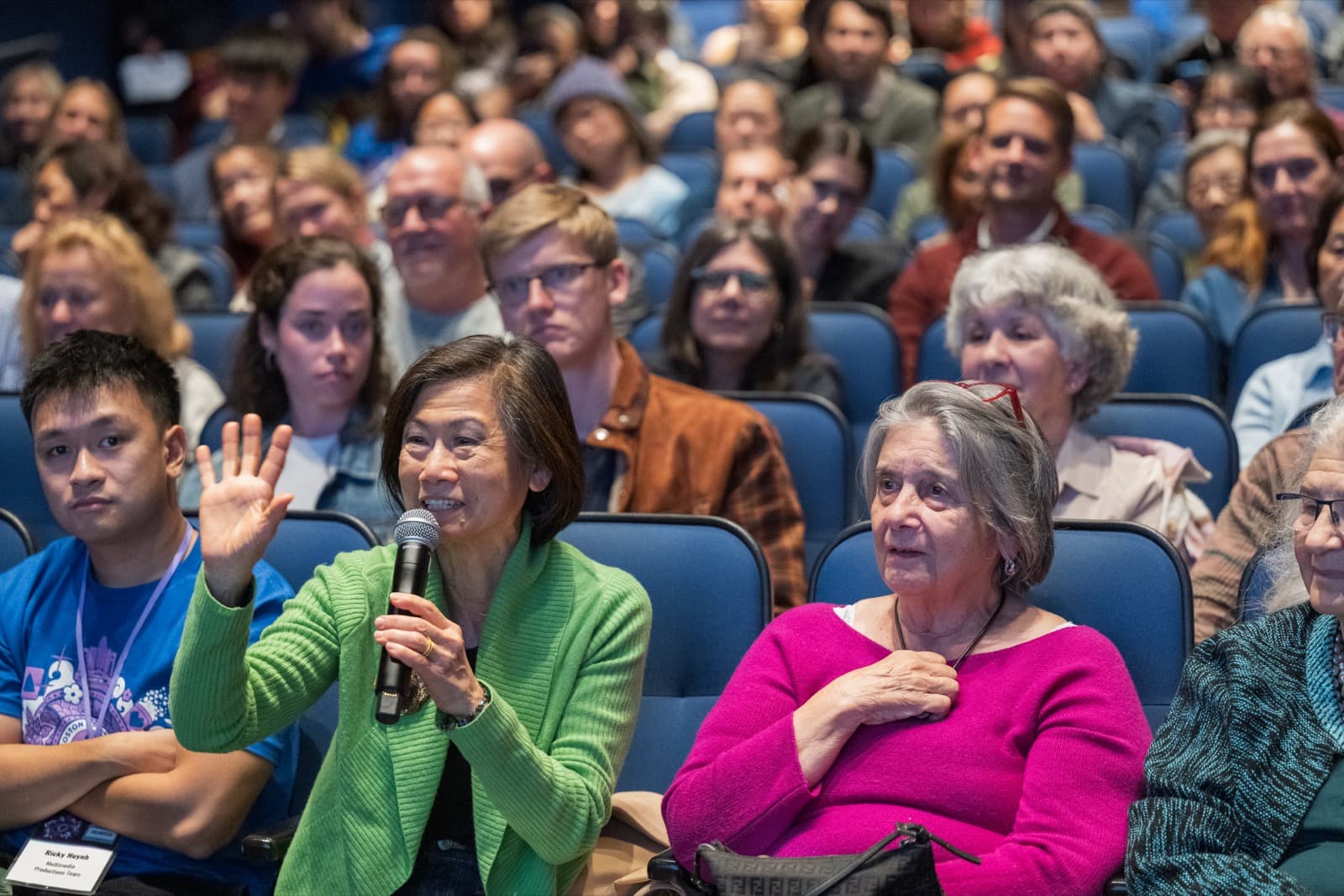 Audience member speaking into a microphone, hand raised mid-sentence, a packed theater crowd behind her.