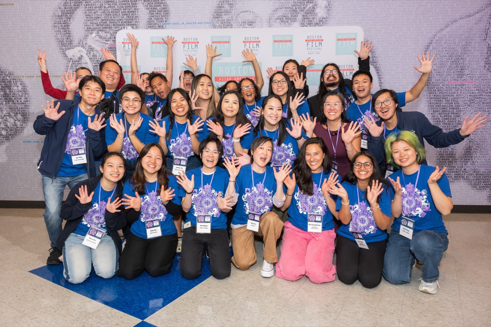 Large group of festival staff and volunteers in matching blue t-shirts, hands raised at the step-and-repeat.