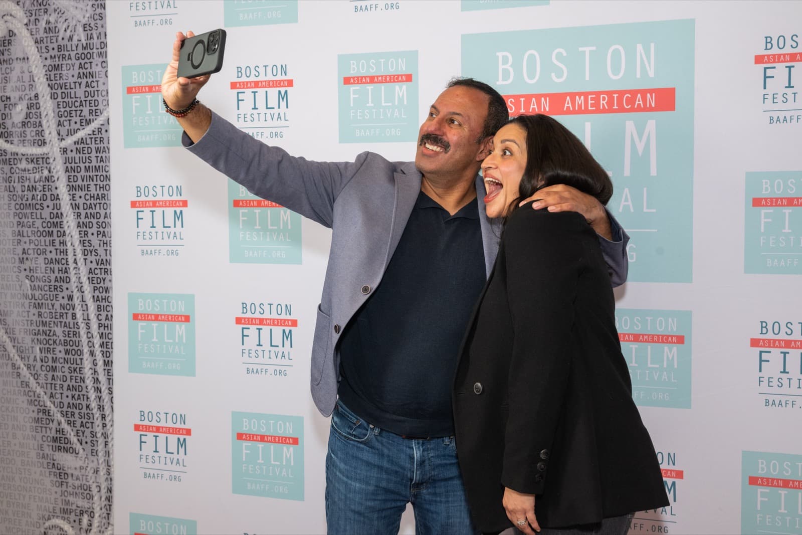 Two people smiling for a selfie together in front of the step-and-repeat backdrop.