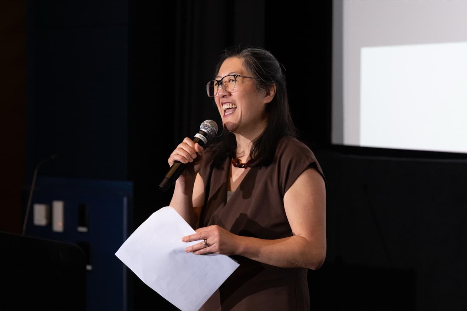 Woman beaming mid-speech into a microphone, notes in hand, a presentation screen behind her.