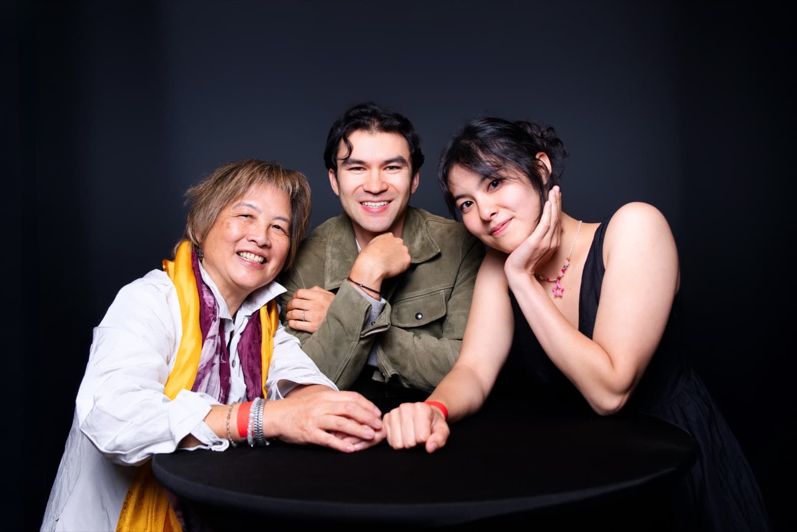 Three people leaning together over a round table in a studio portrait, warm expressions all around.
