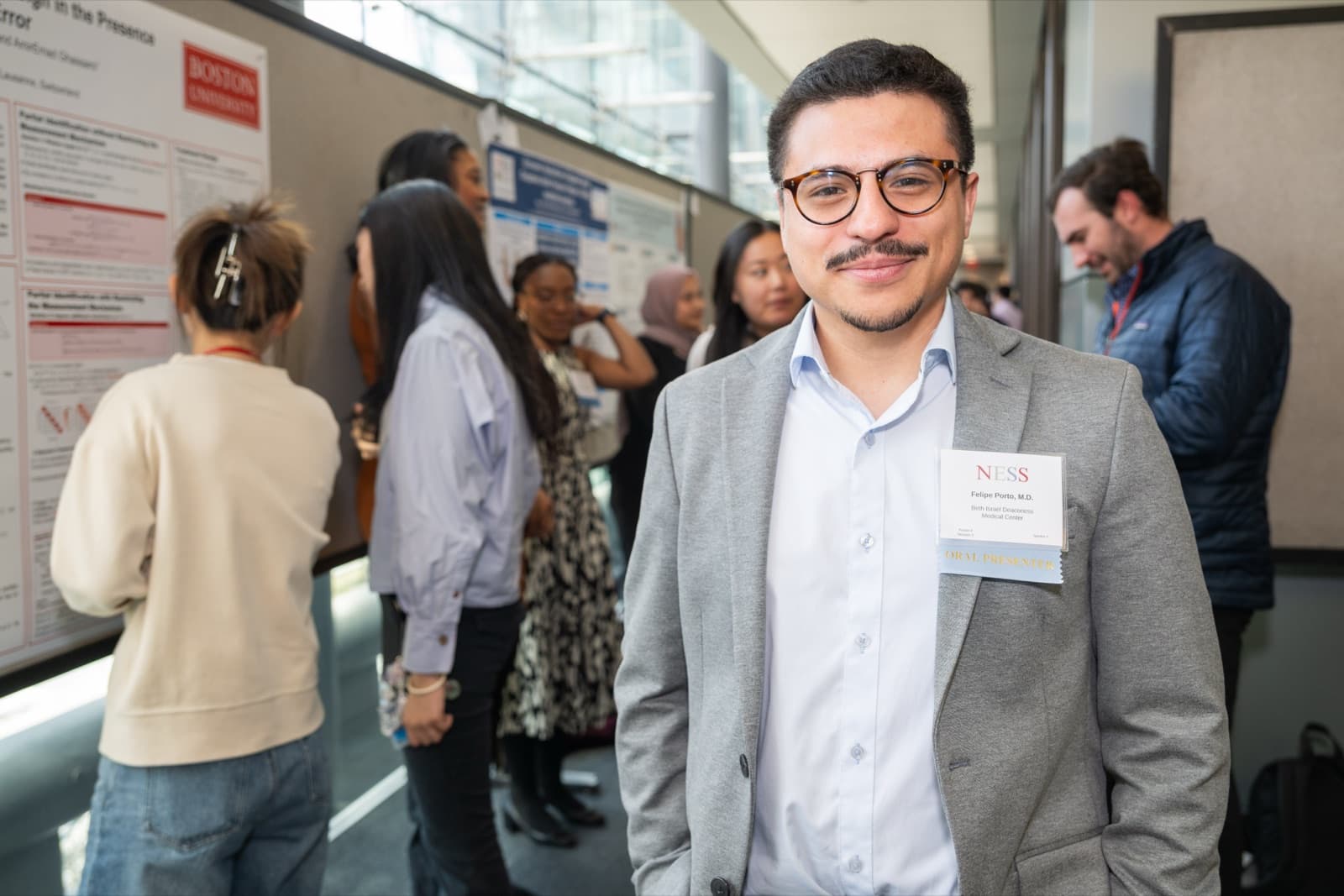 Man in glasses and a mustache, delighted expression, other attendees viewing research posters behind him in a glass hallway.