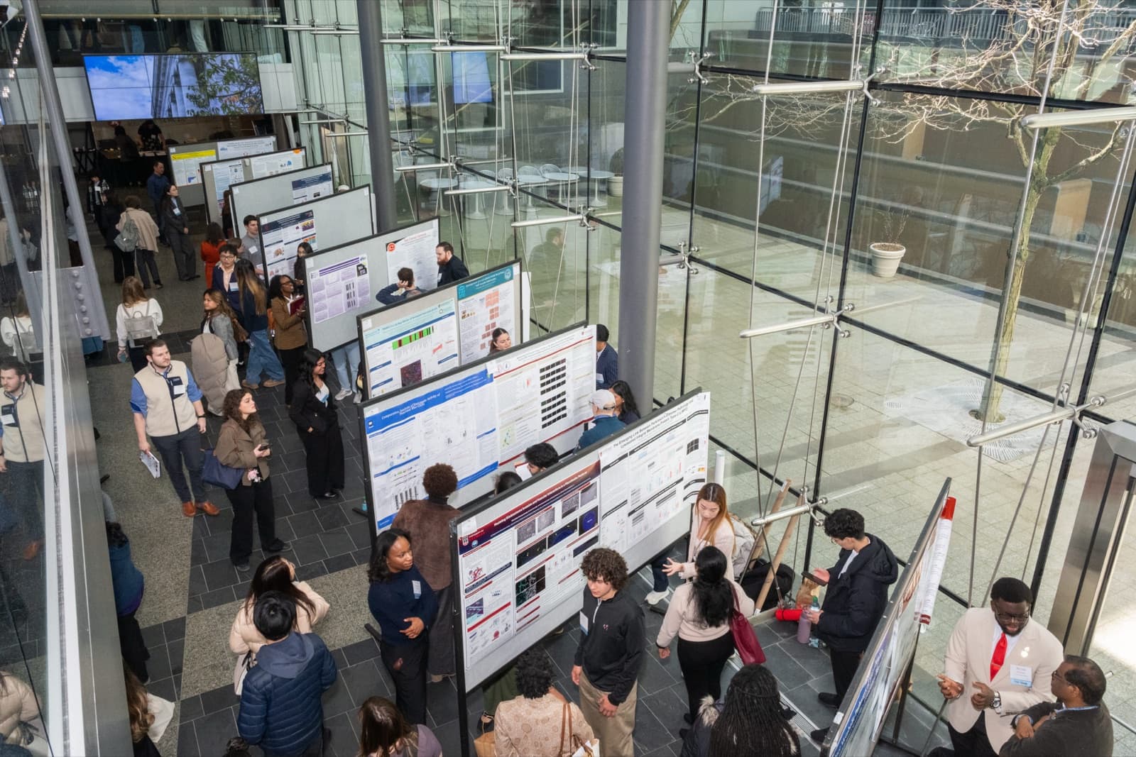 Aerial view of a crowded research poster session, dozens of attendees browsing rows of presentation boards in a glass atrium.