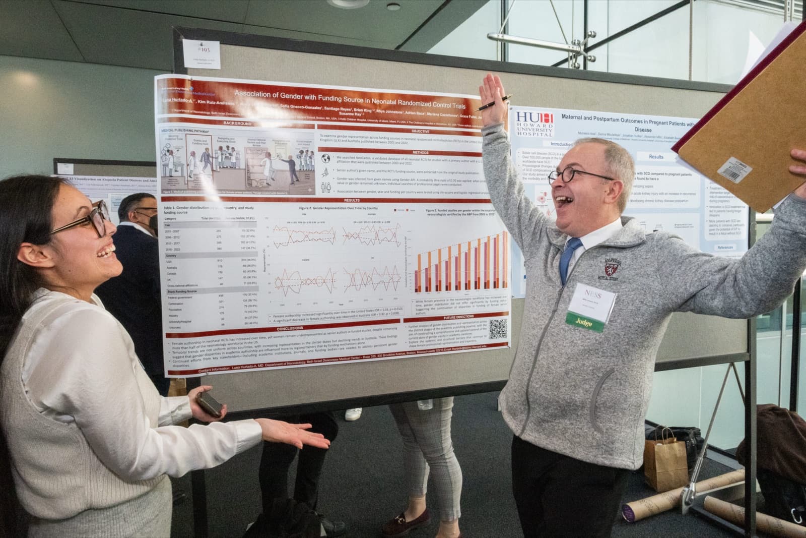 Man raising his fist triumphantly, holding papers up, a woman beaming beside him, in front of research posters.