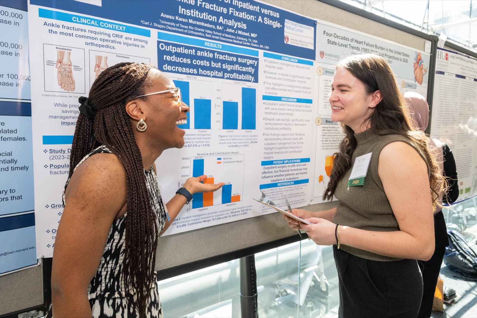 Two women talking face to face, one laughing and gesturing at a research poster with bar charts.