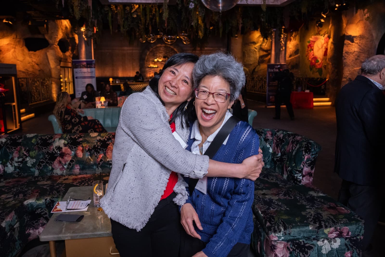 Two women embracing and laughing at the camera, one in a gray cardigan and one in blue knit, warm-lit event space behind.
