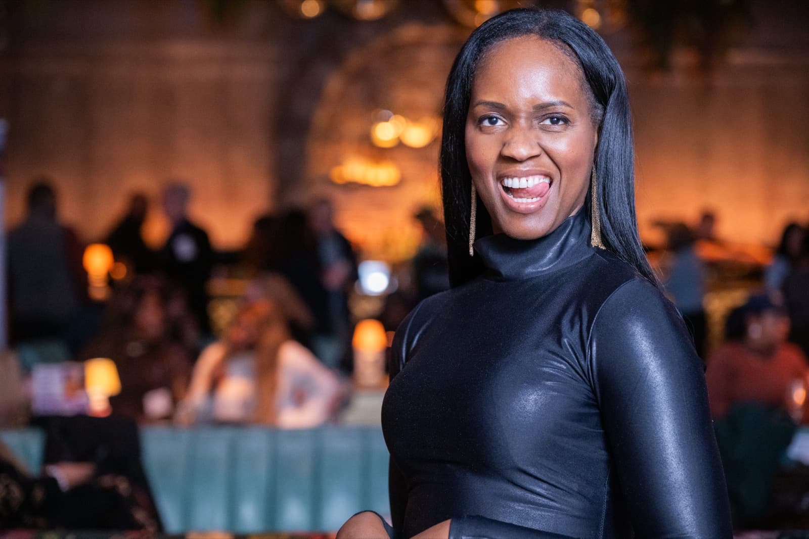 Woman with long black hair laughing brightly in a black high-neck top and gold drop earrings, guests blurred behind her.