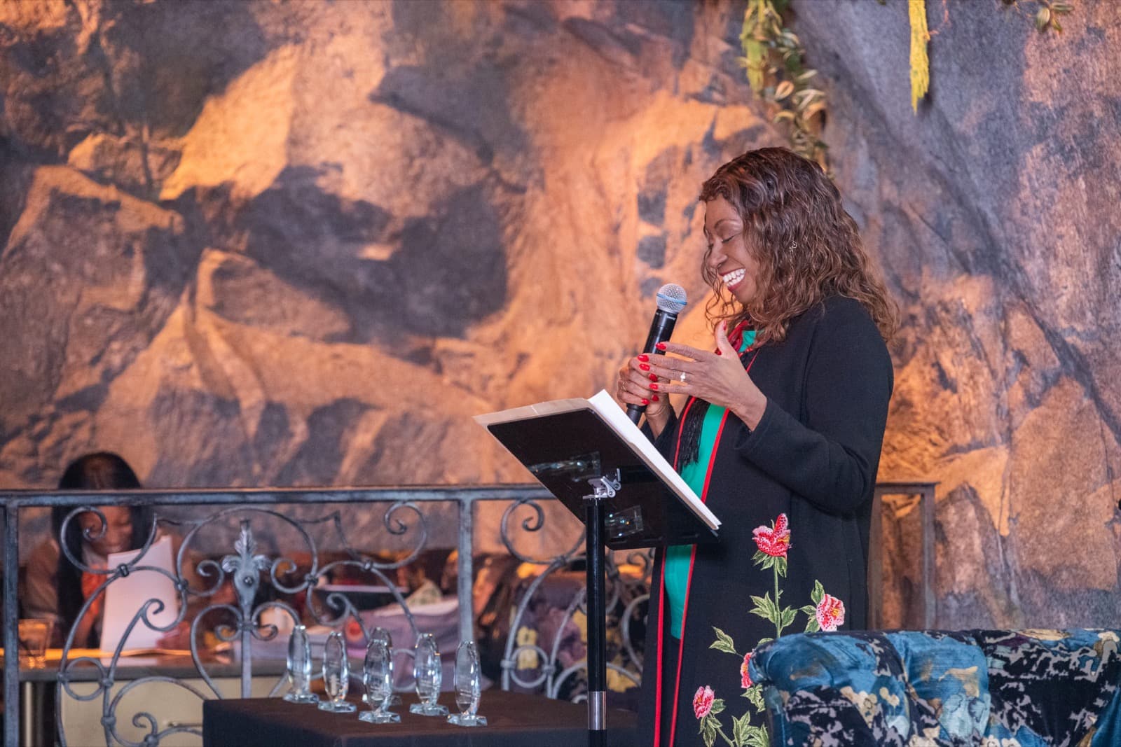 Woman speaking into a handheld microphone at a slim podium, floral-embroidered jacket, glass awards lined up in front.