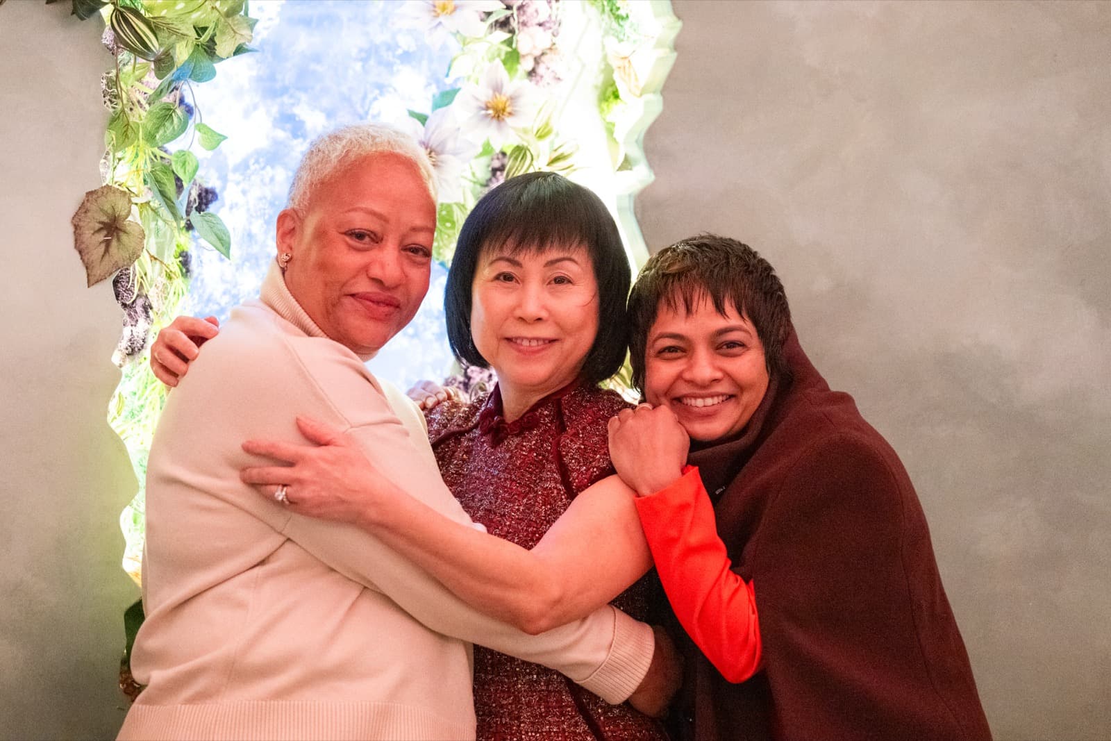 Three women posed close together in front of a floral backdrop, arms around each other, all smiling at the camera.