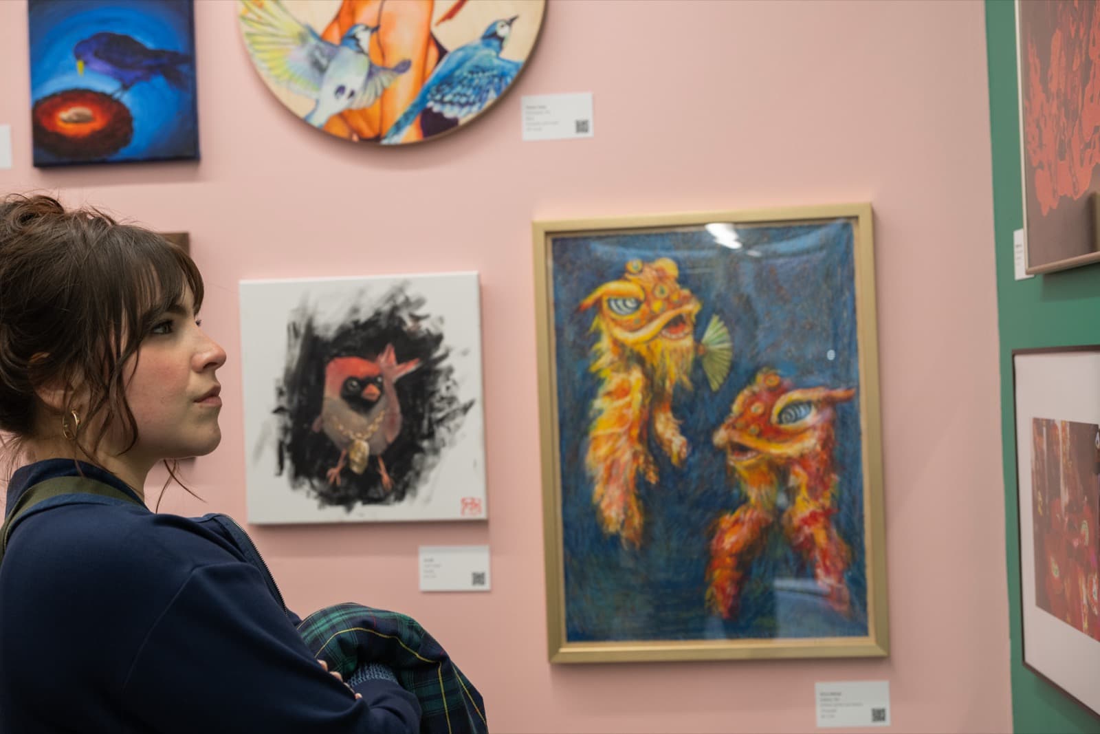 Gallery visitor studying a wall of framed bird paintings and a colorful lion-dance mask illustration.