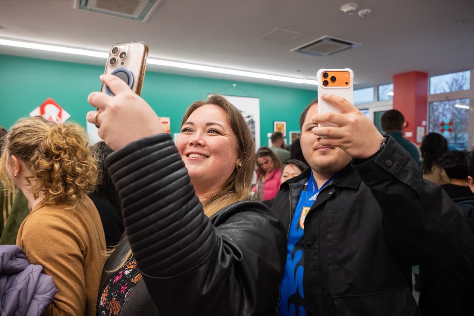 Two people smiling as they take a selfie together at a crowded opening, vivid artwork covering the wall behind them.