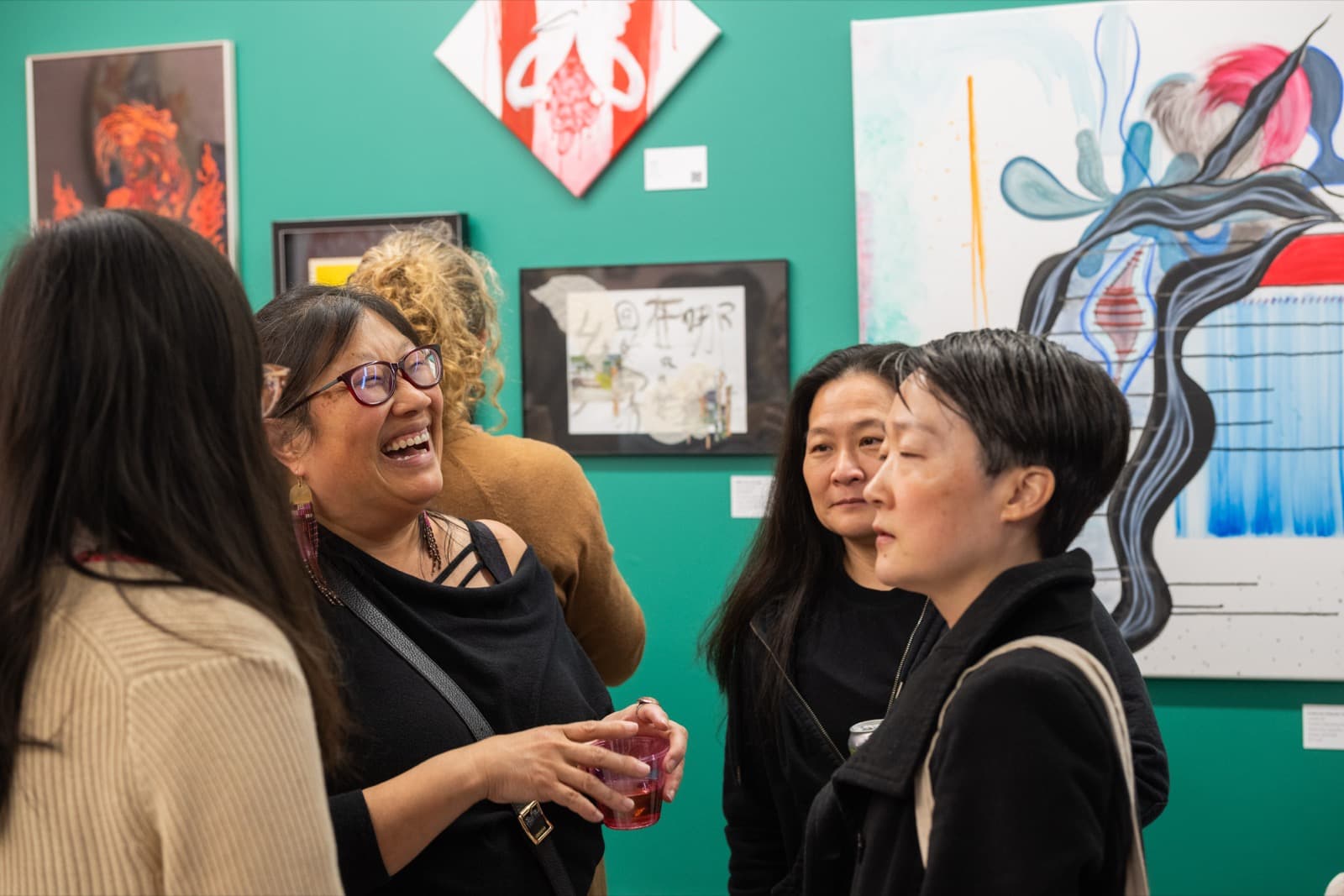 Group of people laughing and chatting in front of vibrant mixed-media artwork on a teal gallery wall.