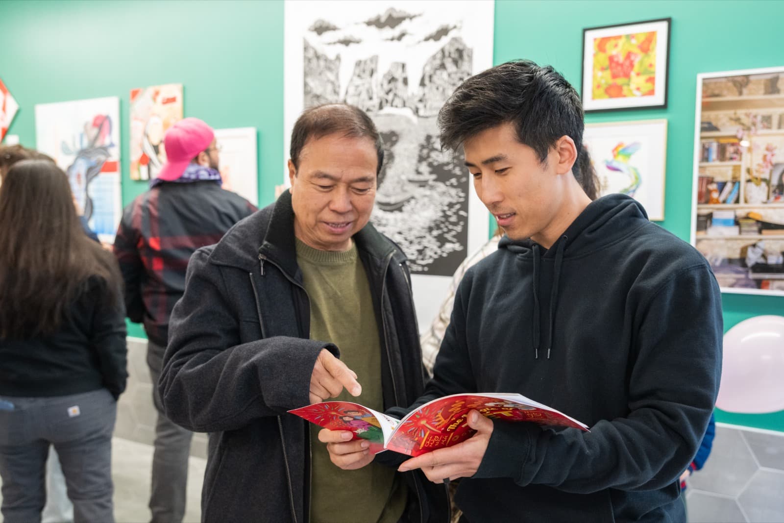 Man and boy looking together at a children's book, a pink balloon and framed ink drawings nearby.