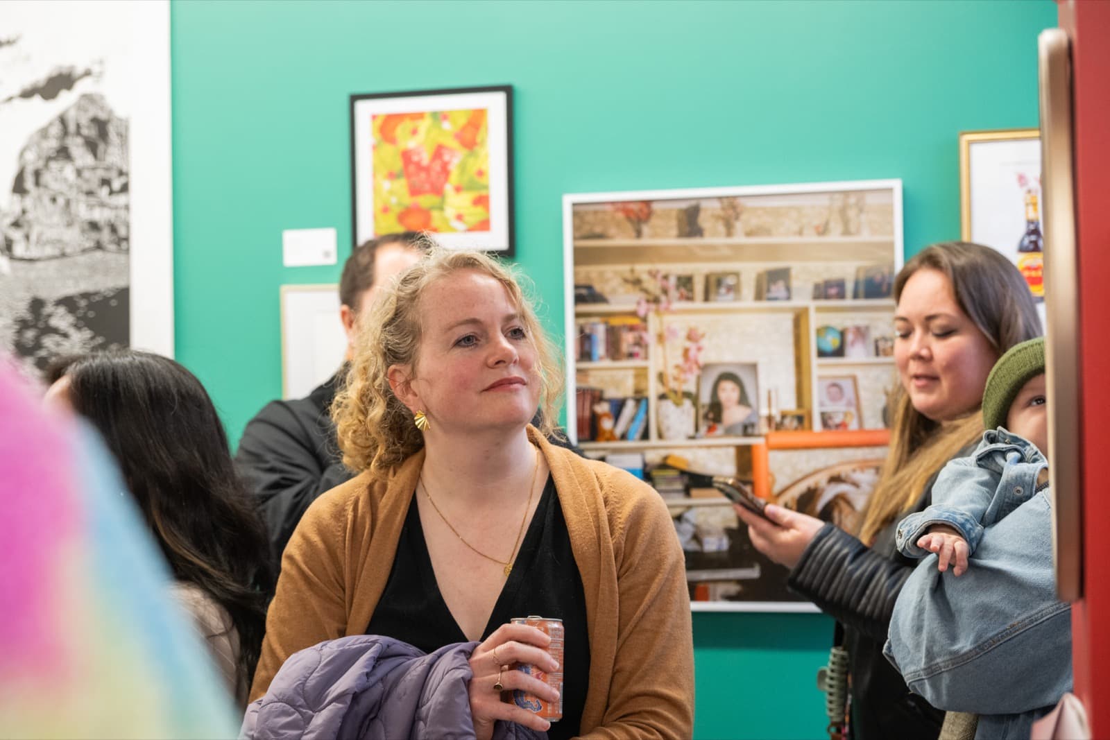 Woman holding a baby, looking at her phone beside a visitor admiring a photo collage of a decorated bookshelf.