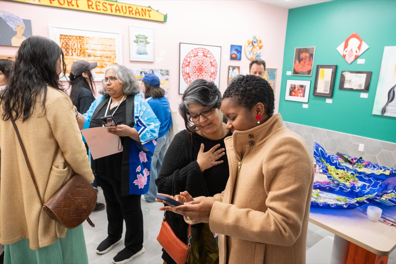 Group of visitors chatting and looking at their phones in front of framed artwork near a restaurant sign.
