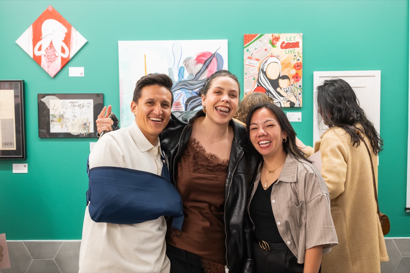 Three people laughing together for a photo, one with his arm in a sling, in front of colorful protest-art paintings.