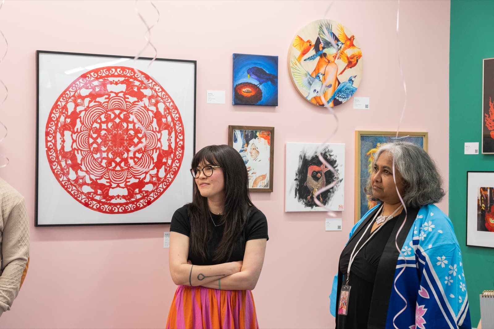 Two women looking up at a gallery of bird paintings and a large red paper-cut mandala design, streamers in the foreground.