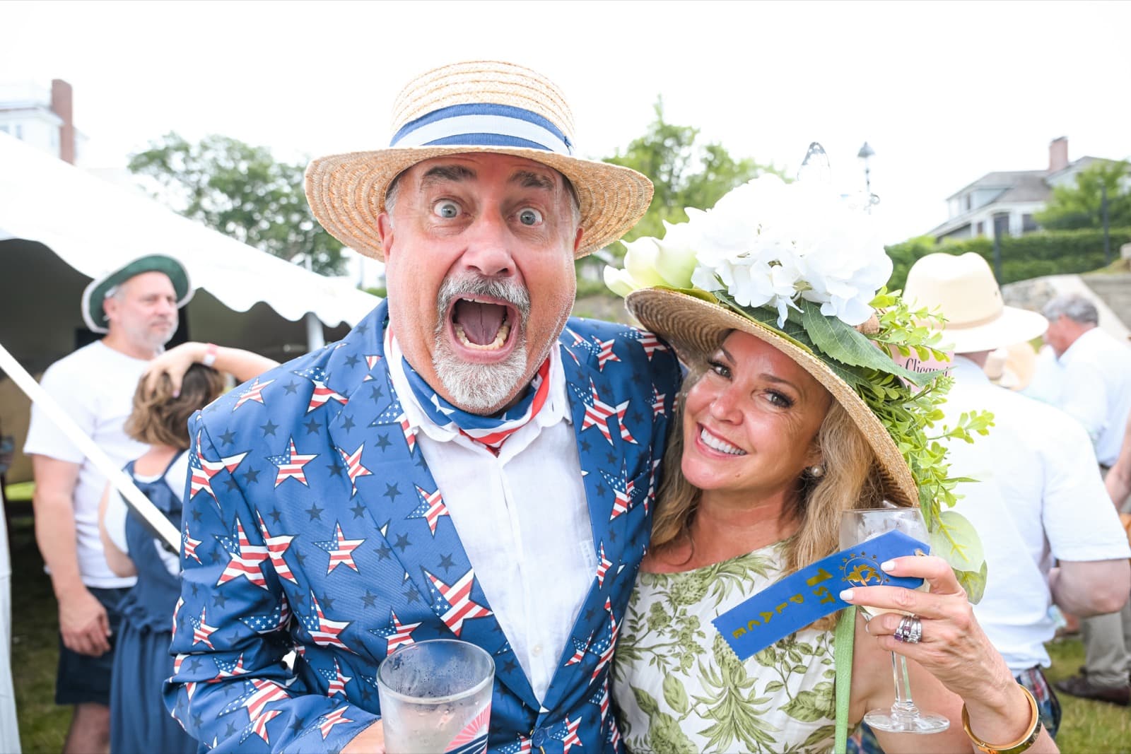 Man in a star-spangled blazer and boater hat gasping with excitement, arm around a woman in a flowered sun hat and blue-ribbon rosette.