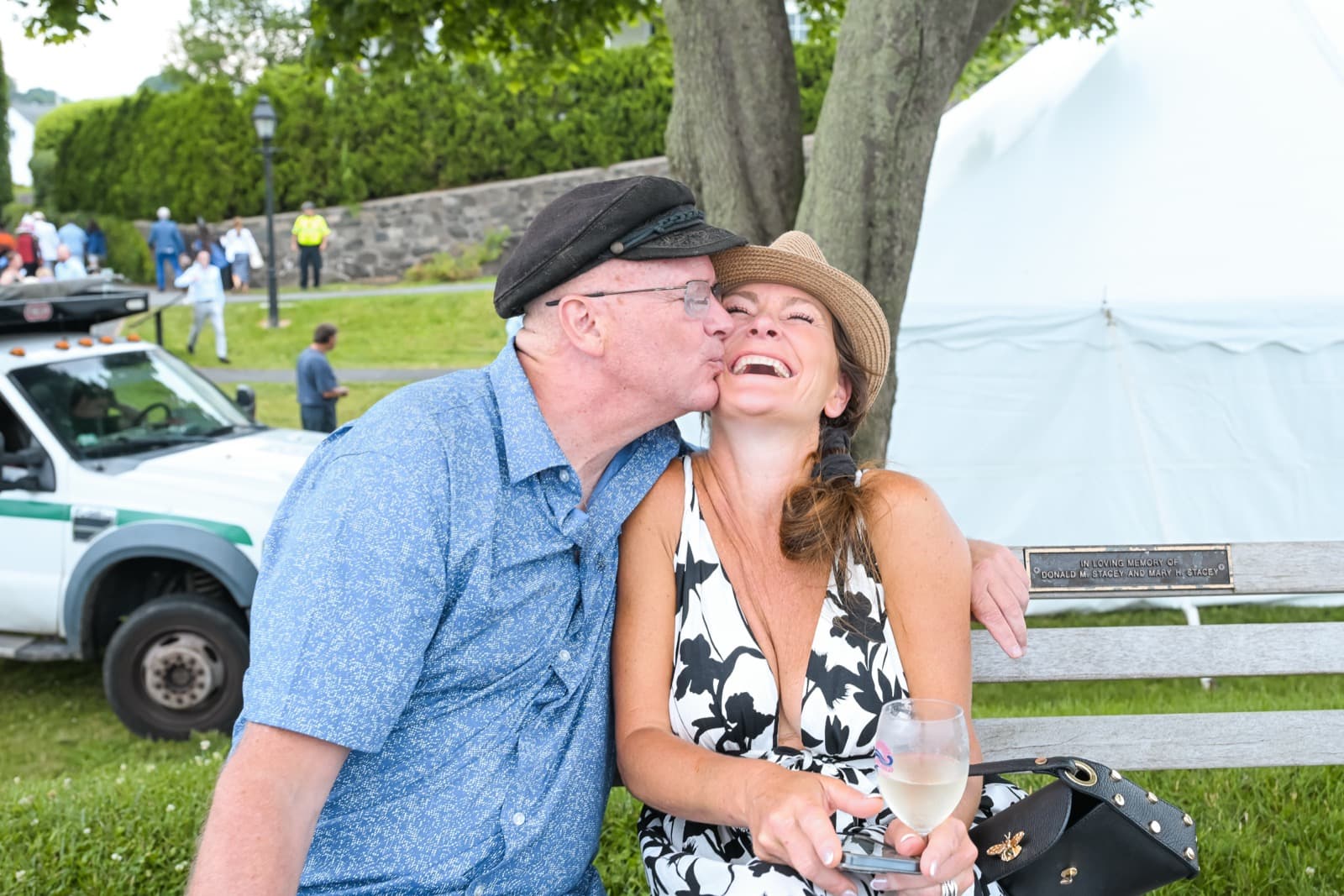 Man in a flat cap kissing a laughing woman on the cheek on a park bench, a festival tent and parked truck behind them.