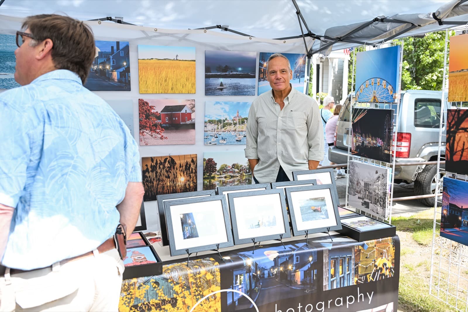 Landscape photographer standing behind his framed prints of harbor scenes at his booth, a visitor browsing.