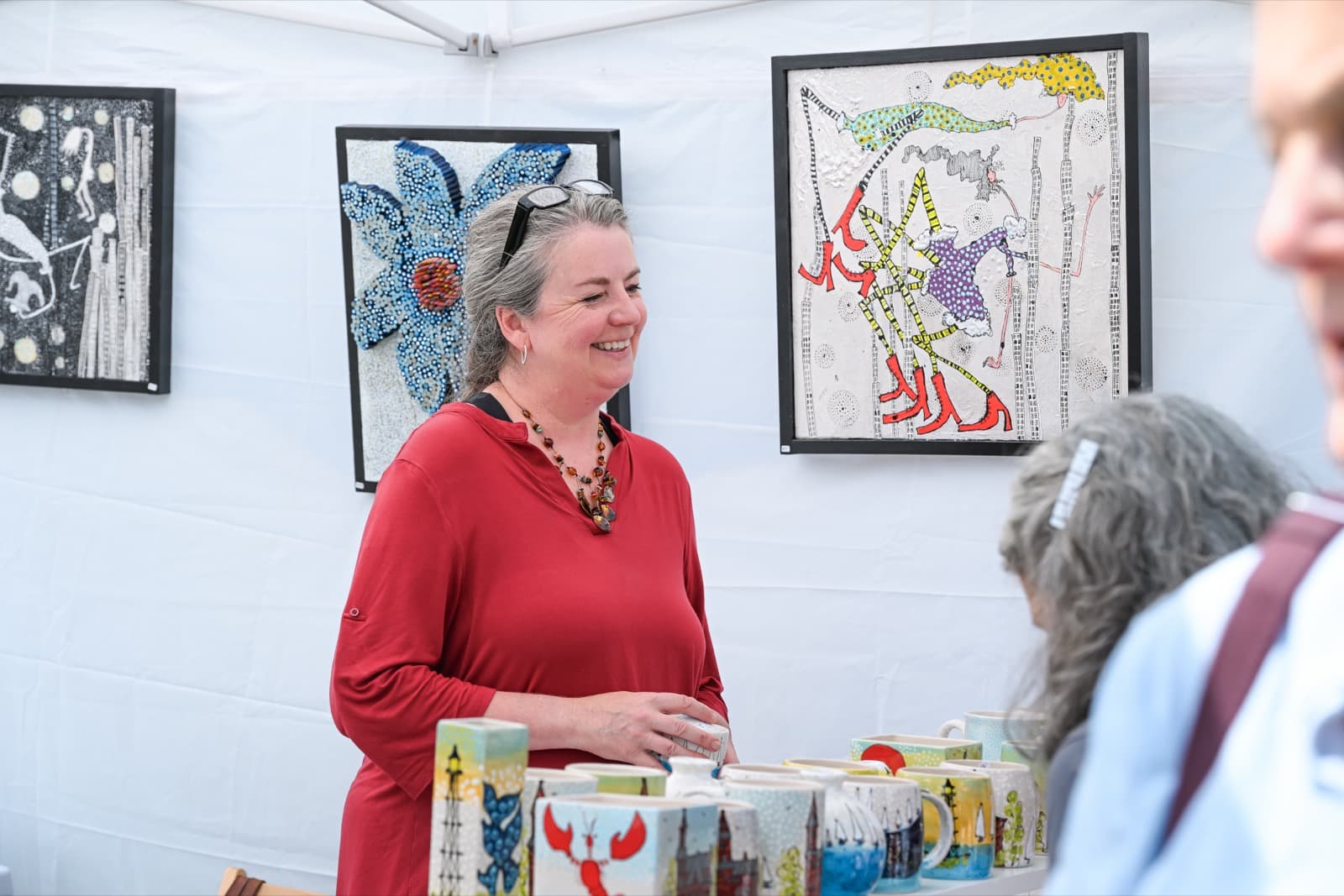 Ceramic artist in a red dress smiling in her booth, surrounded by colorful painted mugs and mixed-media artwork.