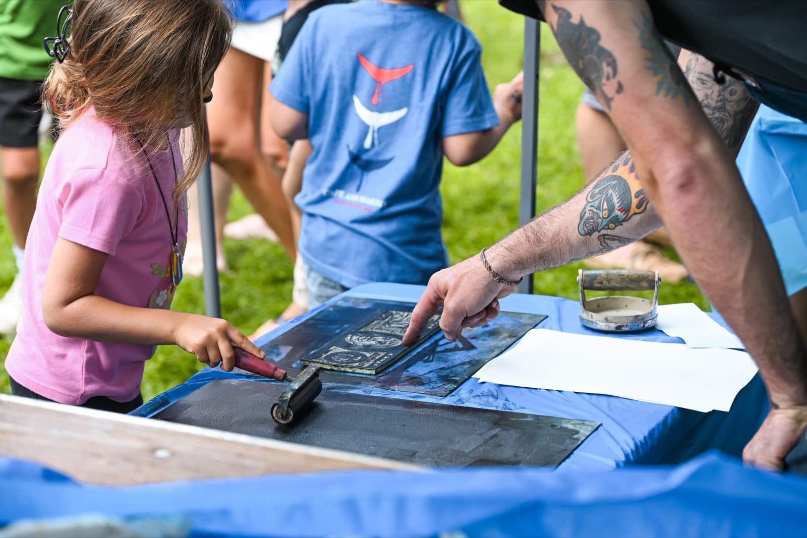 A child's and a tattooed artist's hands working a linocut print together at a printmaking demo table.